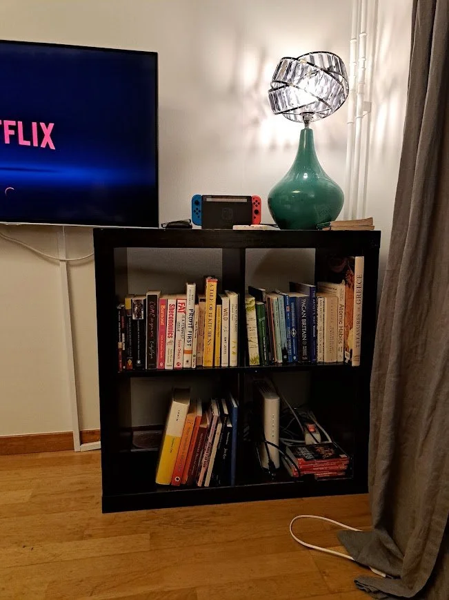 A dark, square bookshelf on a light wood floor. It's filled with books and on top is a large, teal lamp
