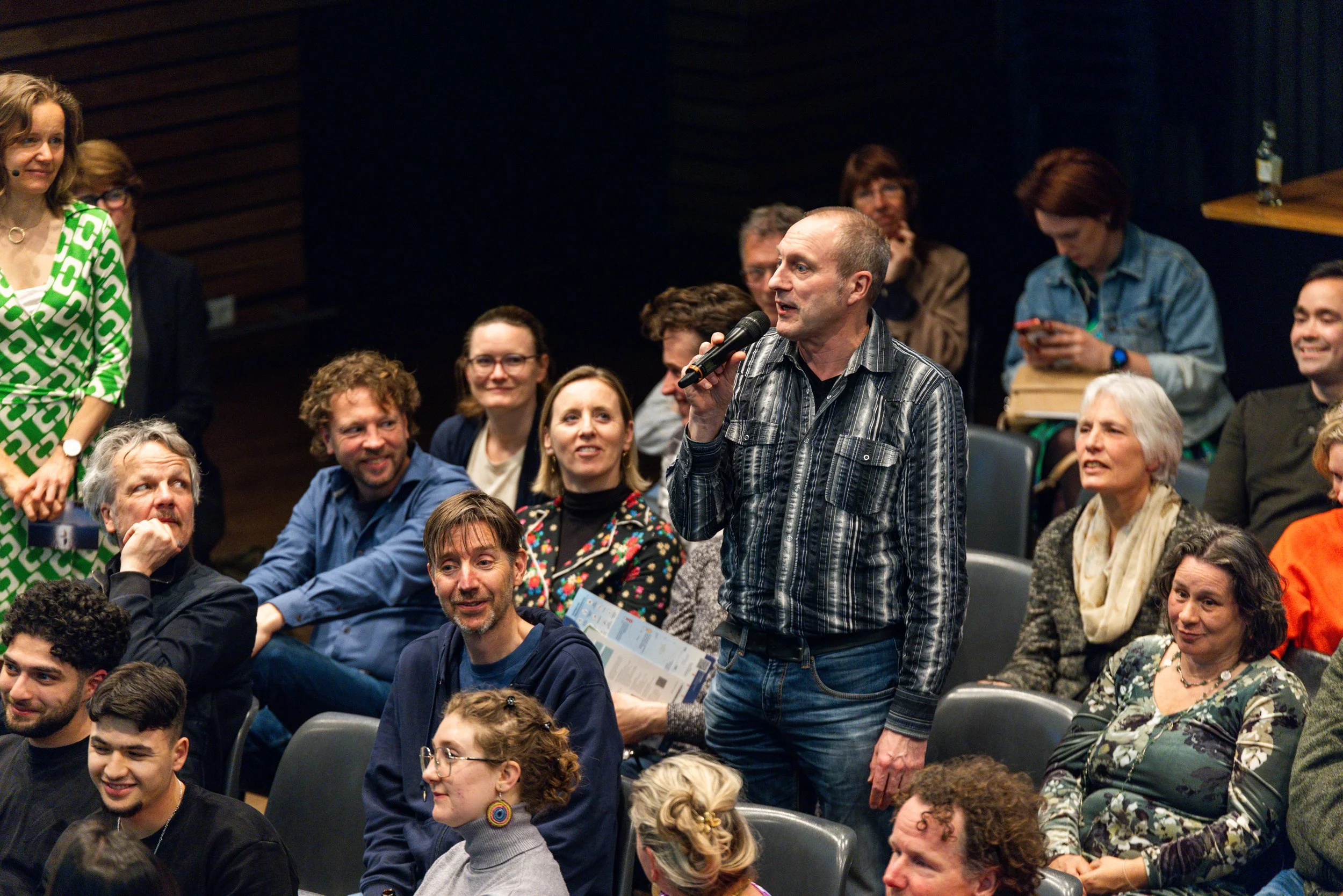 A man with a striped shirt holding a microphone speaks to an audience in a conference or seminar setting, with several people sitting and standing around him.