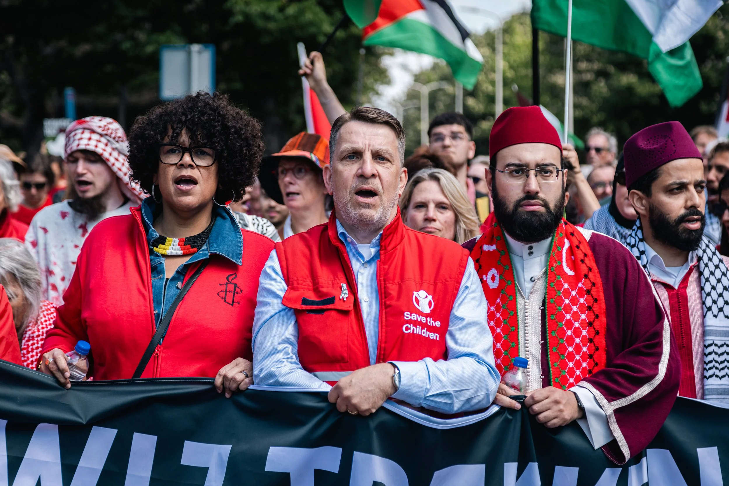 A diverse group of protesters at a demonstration, with some waving flags and holding a banner, wearing red and traditional clothing, participating in a protest or rally.