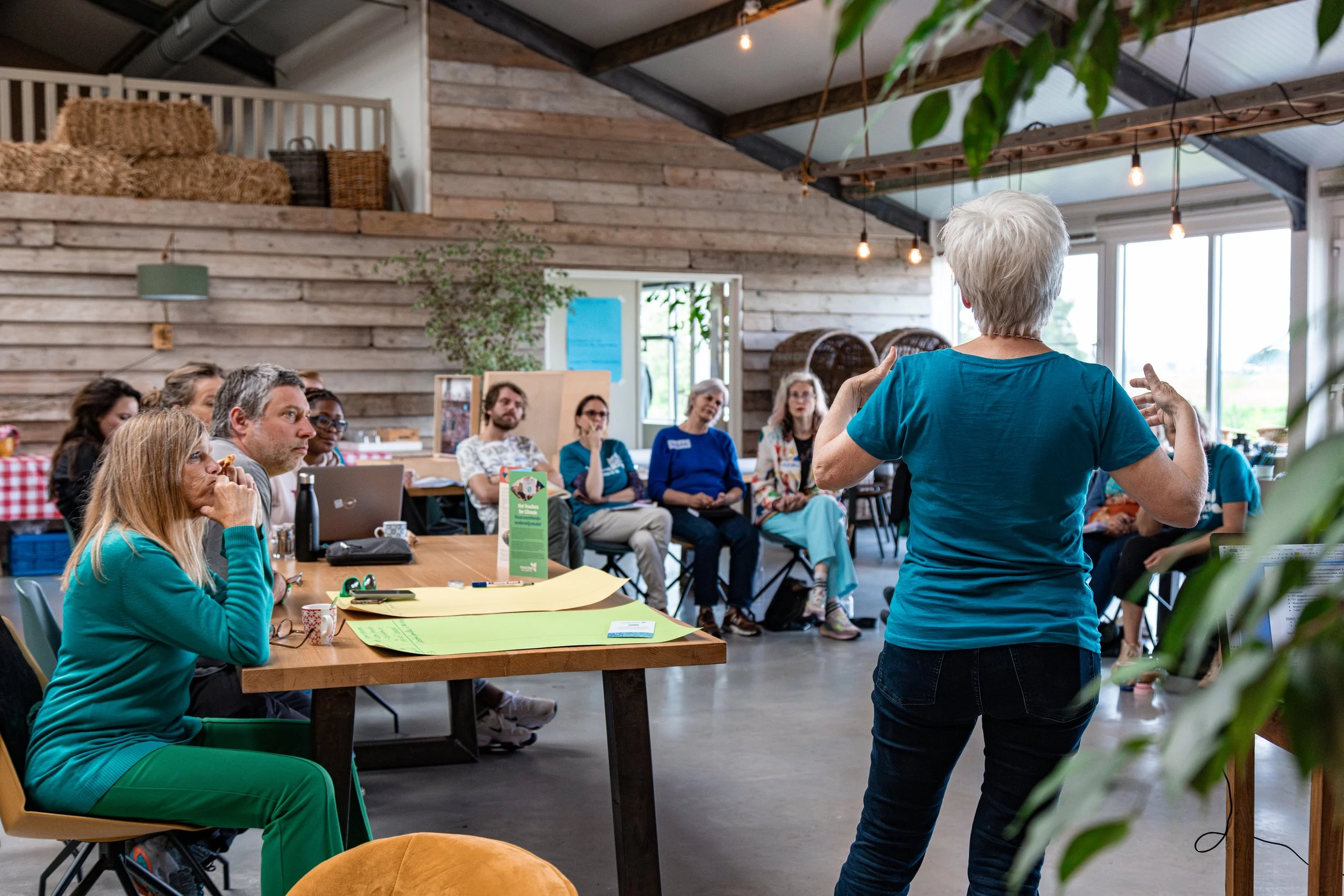 A woman with short gray hair giving a presentation to a group of people seated around a table in a rustic, well-lit room with wooden walls and large windows.