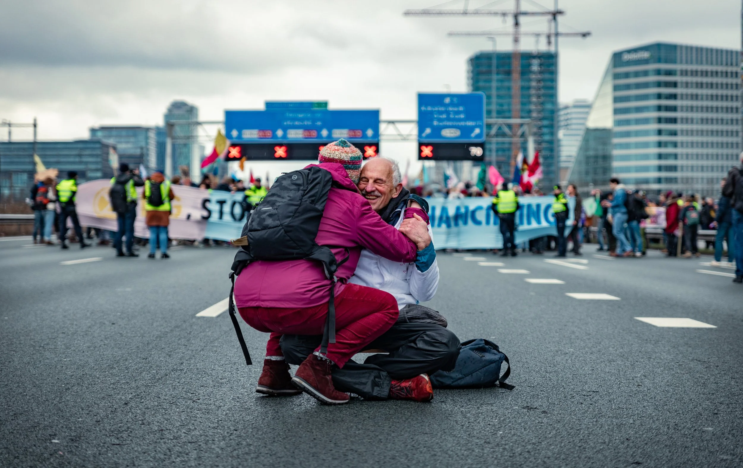 A woman and a man hugging in the middle of a protest on a city street, with protesters and banners in the background.