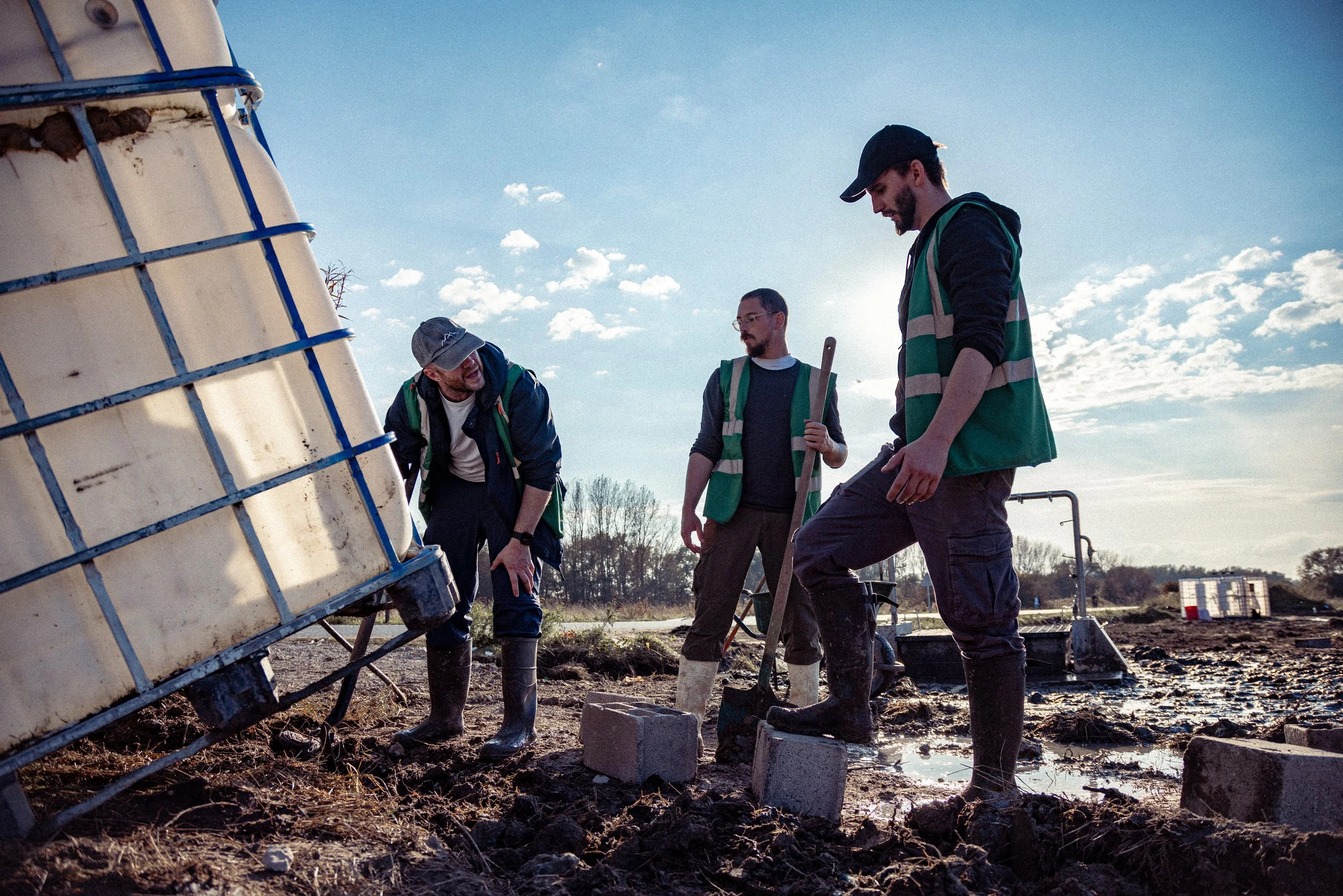 Three men working outdoors on a construction or environmental project, wearing safety vests and boots, with a large water container tilted on the ground and construction equipment in the background under a cloudy sky.