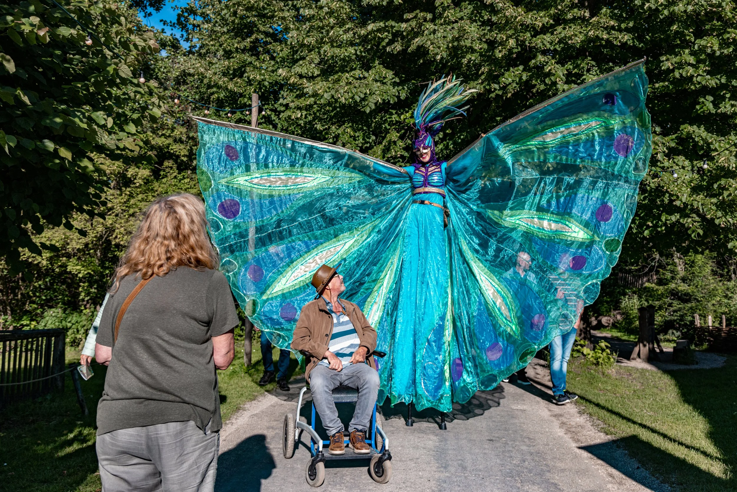 A performer dressed as a butterfly with large, blue, green, and purple wings, standing outdoors as people observe this art installation.