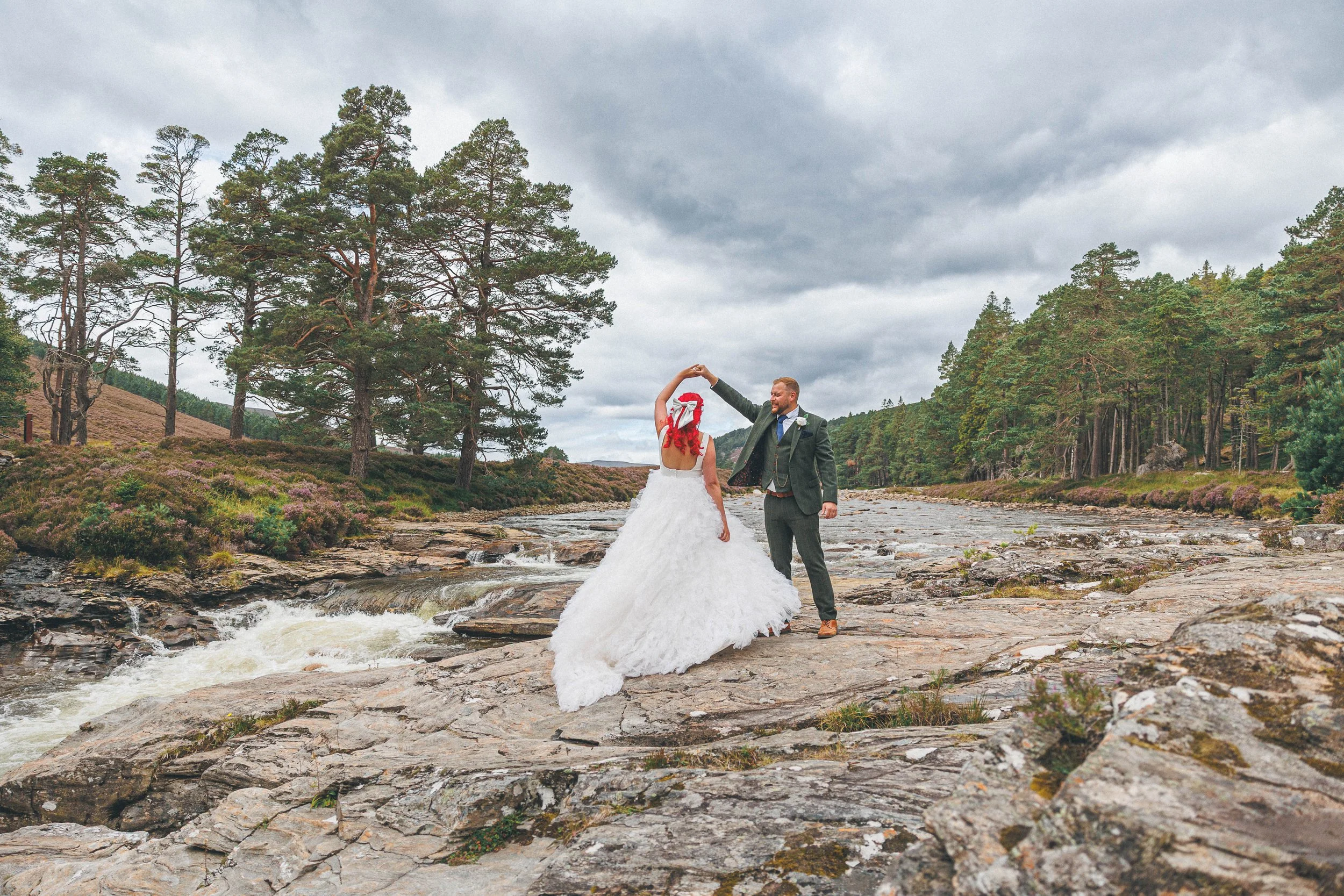A bride and groom dancing on rocks in a river surrounded by trees and cloudy sky.