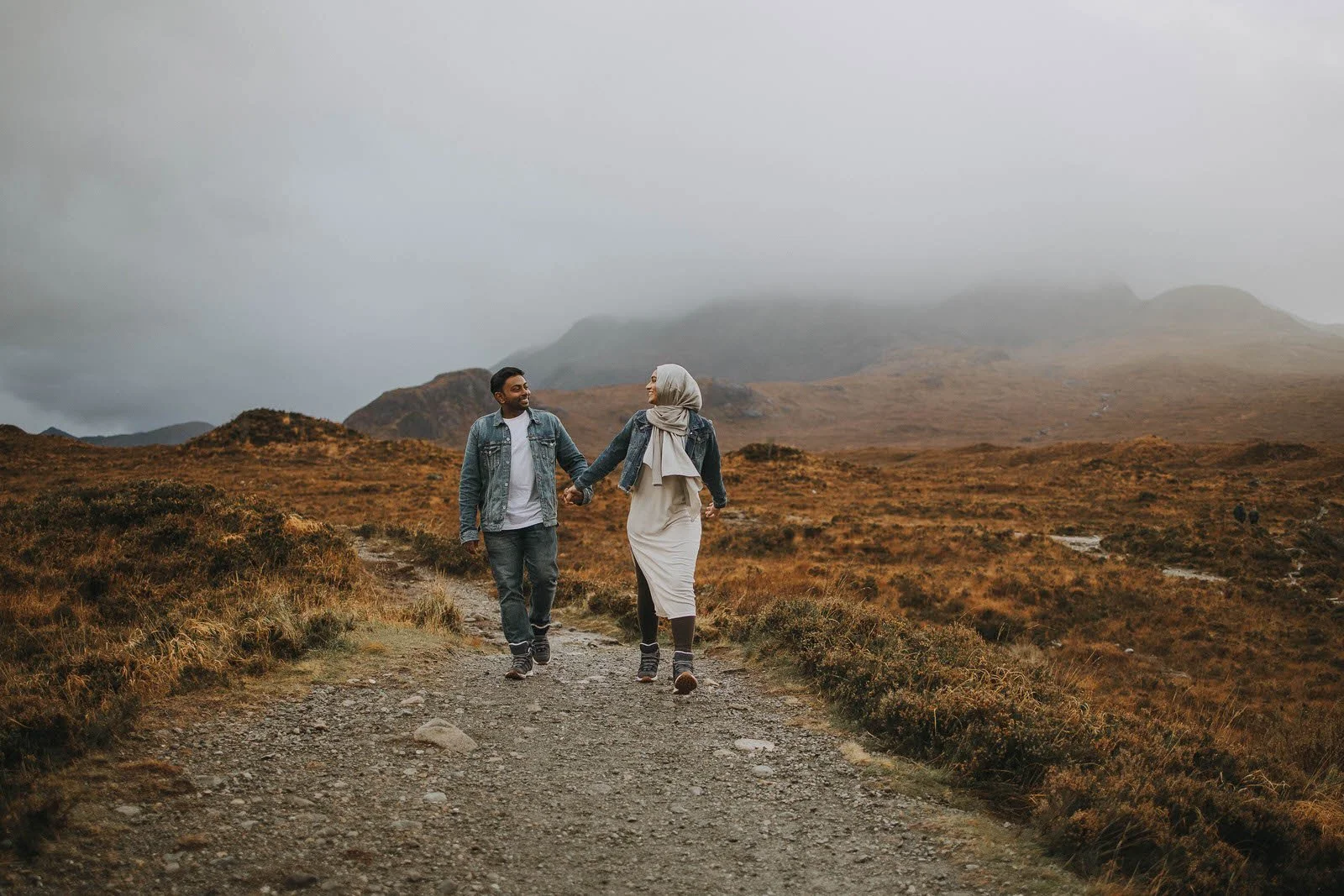 A couple walking hand-in-hand on a dirt trail through a foggy, mountainous landscape with orange-brown foliage.