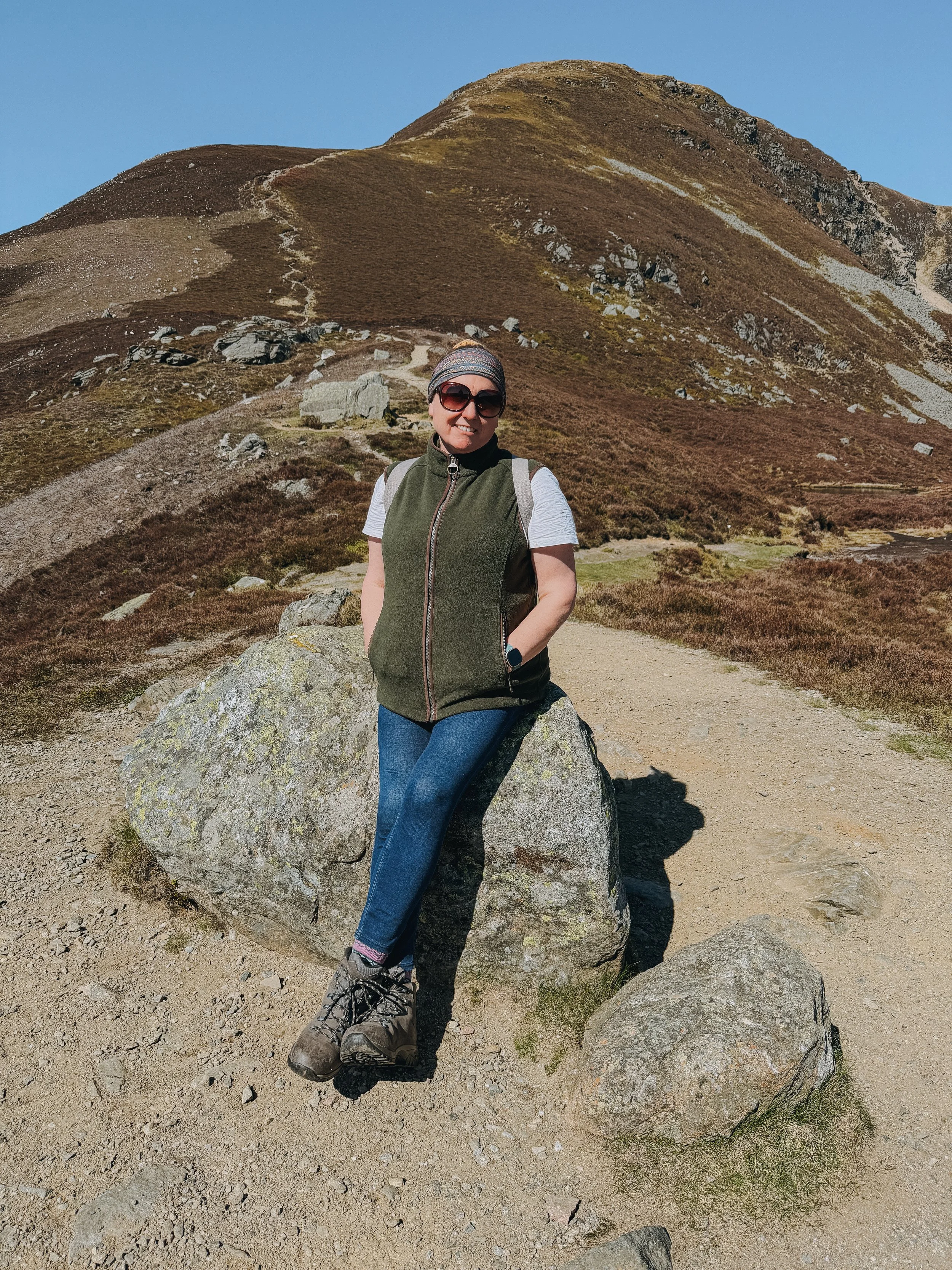 A woman smiling and wearing sunglasses, a headband, a green vest, and hiking boots, standing outdoors in front of a large mountain with a dirt trail.