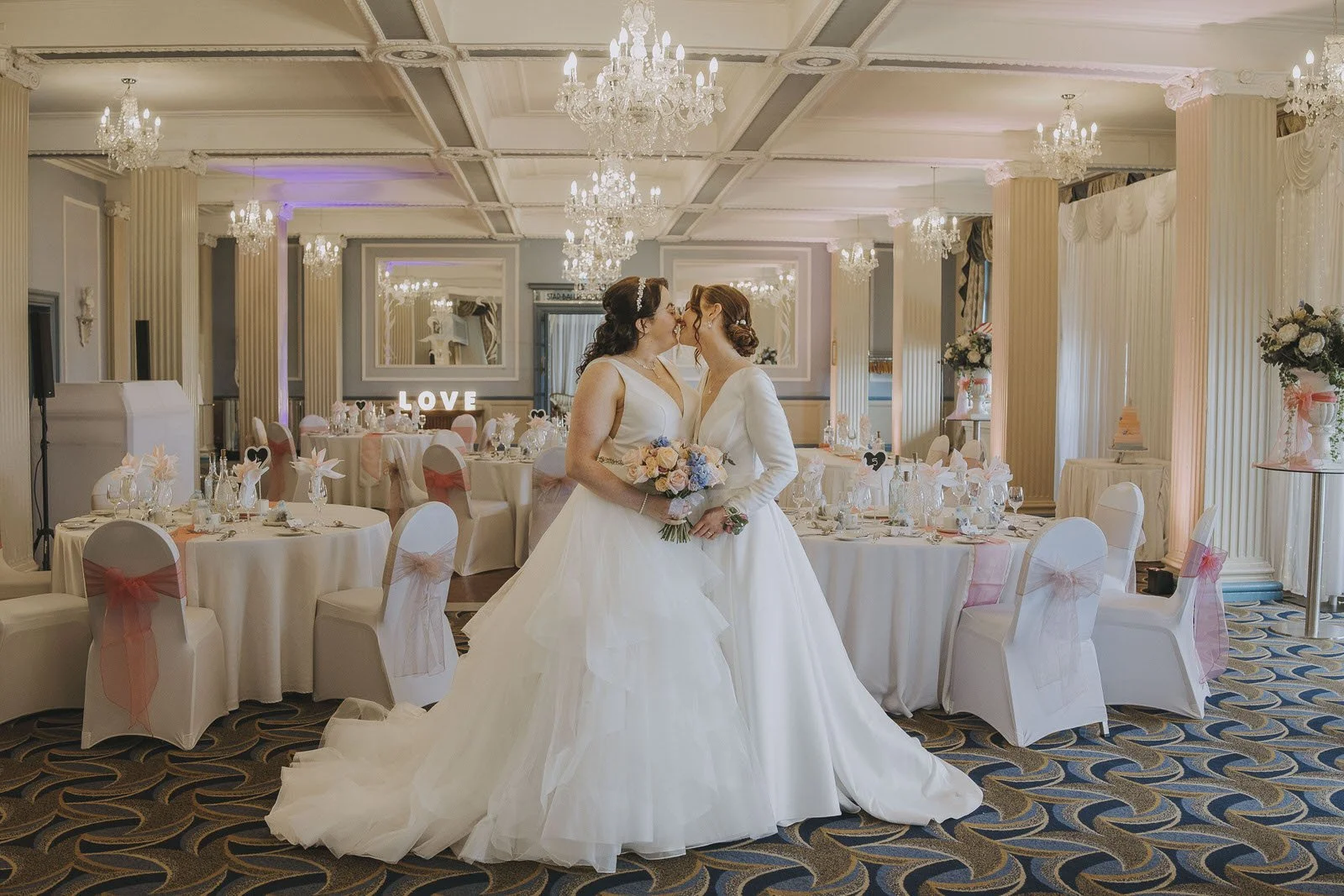 Two brides in wedding dresses, kiss in the beach ballroom Aberdeen in the main room decorated with elegant chandeliers, round tables with white tablecloths, pink accents, and floral centerpieces.