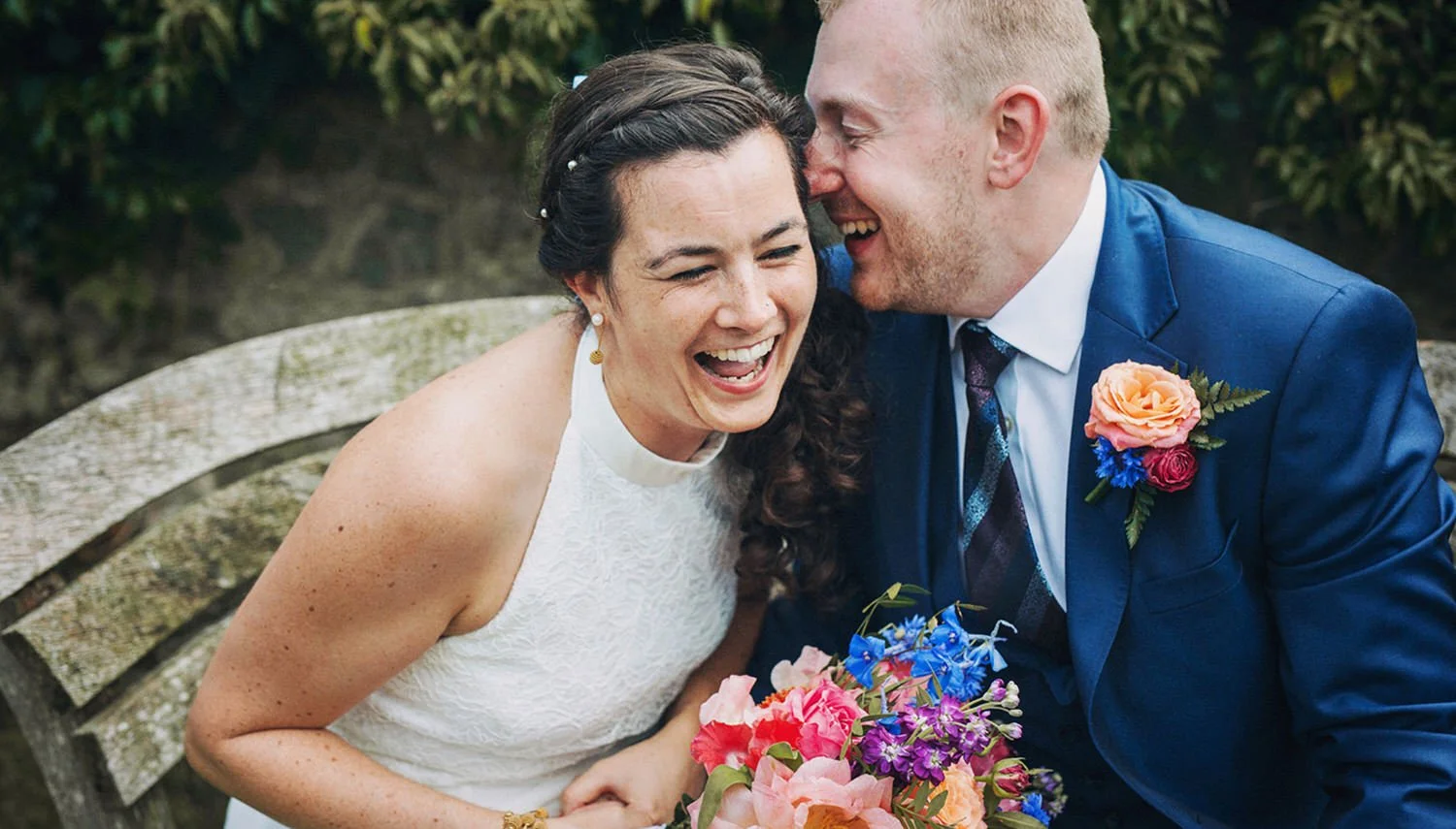 A bride and groom sharing a joyful moment, laughing and leaning close together on a park bench with a bouquet of colorful flowers.