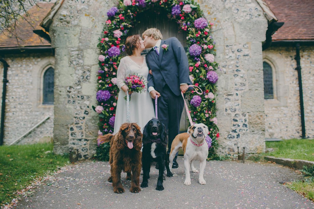 A couple in wedding attire kissing in front of a floral archway with three dogs on leashes.