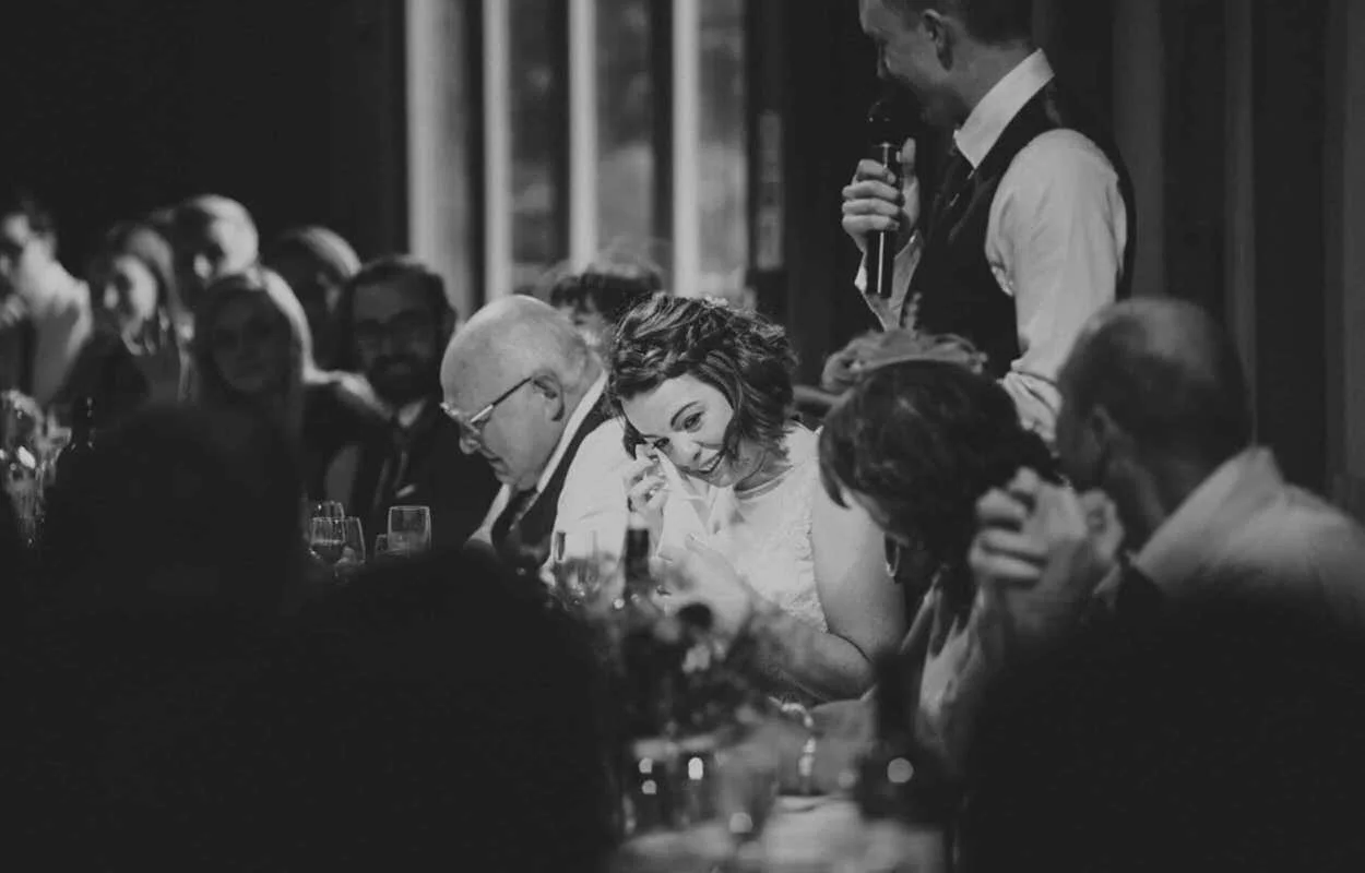 Black and white photo of a woman wiping tears at a formal dinner, surrounded by seated guests, with a waiter standing nearby holding a drink.