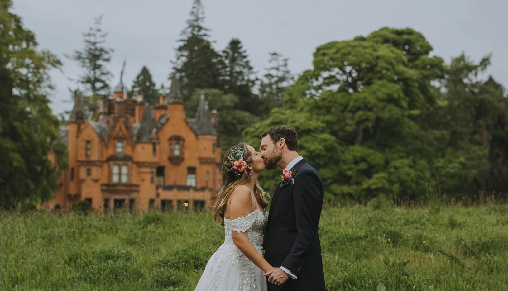 A bride and groom share a kiss outdoors in front of a historic castle, surrounded by lush green trees.