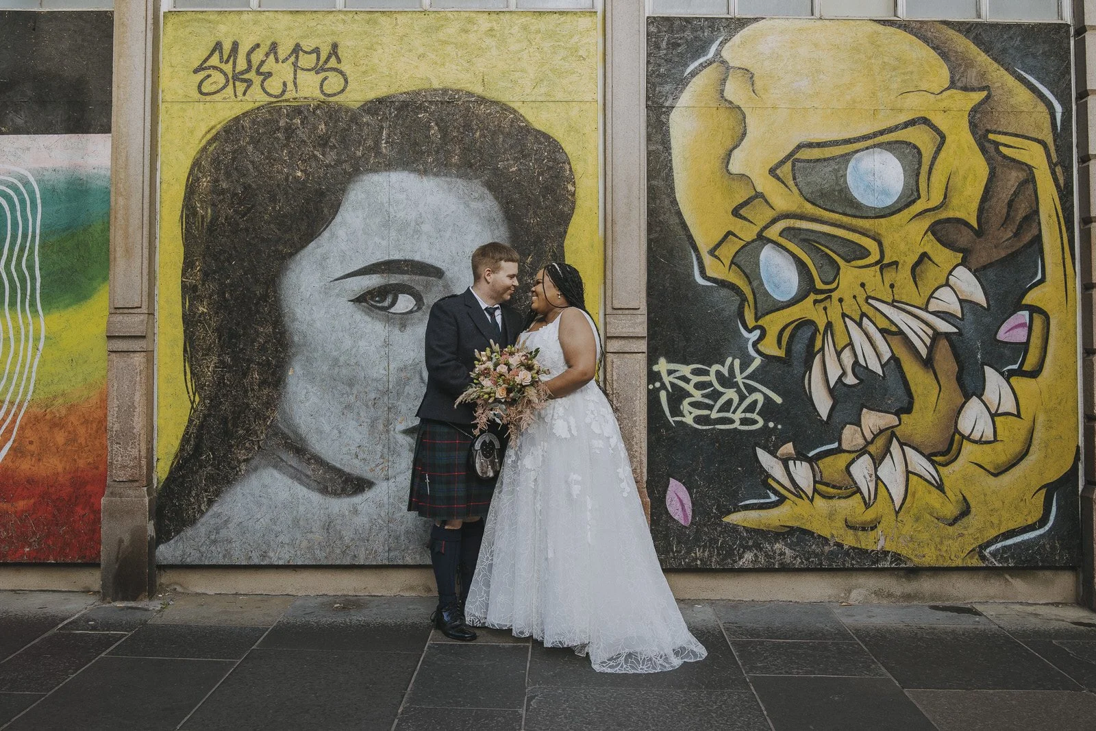 A newlywed couple standing in front of graffiti murals in Aberdeen, with the groom in traditional Scottish attire and the bride in a white wedding dress, holding a bouquet of flowers.