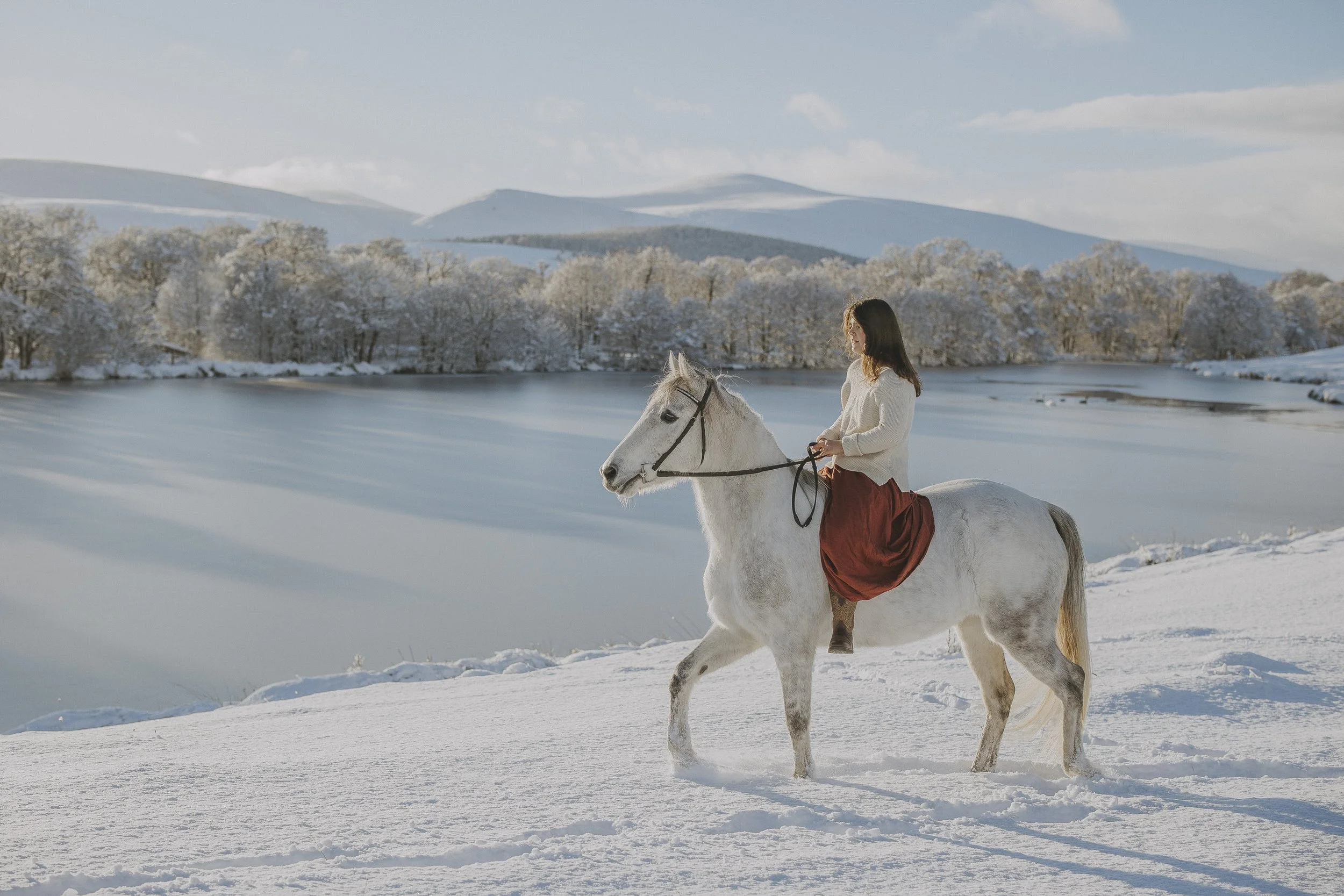 A woman riding a white horse in a snowy landscape with snow-covered trees and mountains in the background.