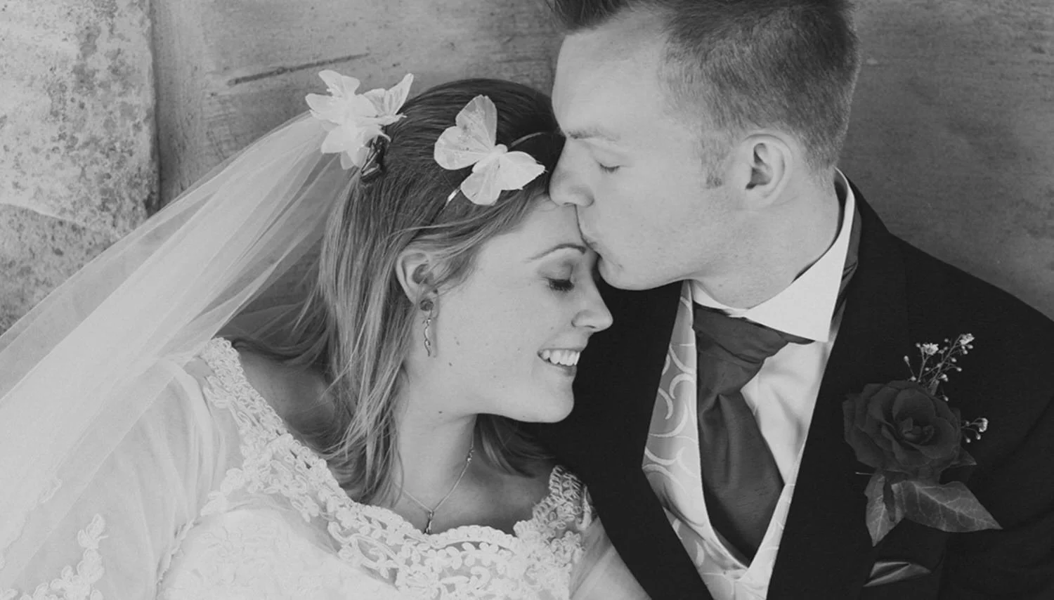 A black-and-white photograph of a bride and groom. The bride is smiling with her eyes closed, resting her head against the groom's forehead. The groom is gently kissing the bride's forehead. She is wearing a lace wedding dress with floral details, and a veil with butterfly hair accessories. He is dressed in a dark suit with a white shirt, patterned tie, and a boutonniere with a rose. They are seated against a stone wall.