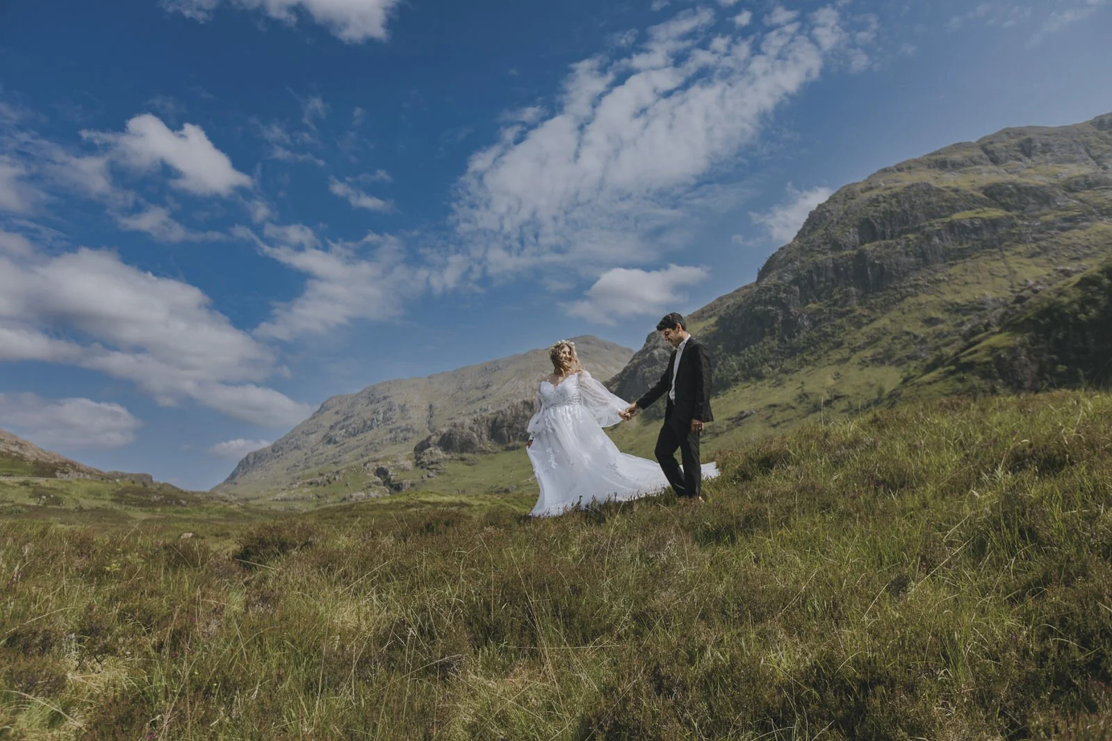 A bride and groom holding hands on a grassy hillside with mountains in the background and a partly cloudy sky.