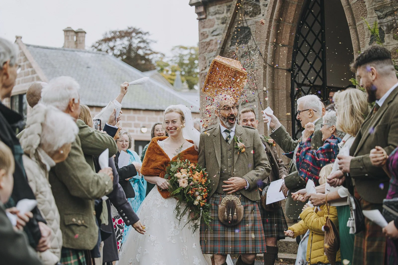 Aberdeenshire photographer captures newlywed couple and guests celebrating outside a church, with the best man throwing confetti and the bride holding a bouquet.