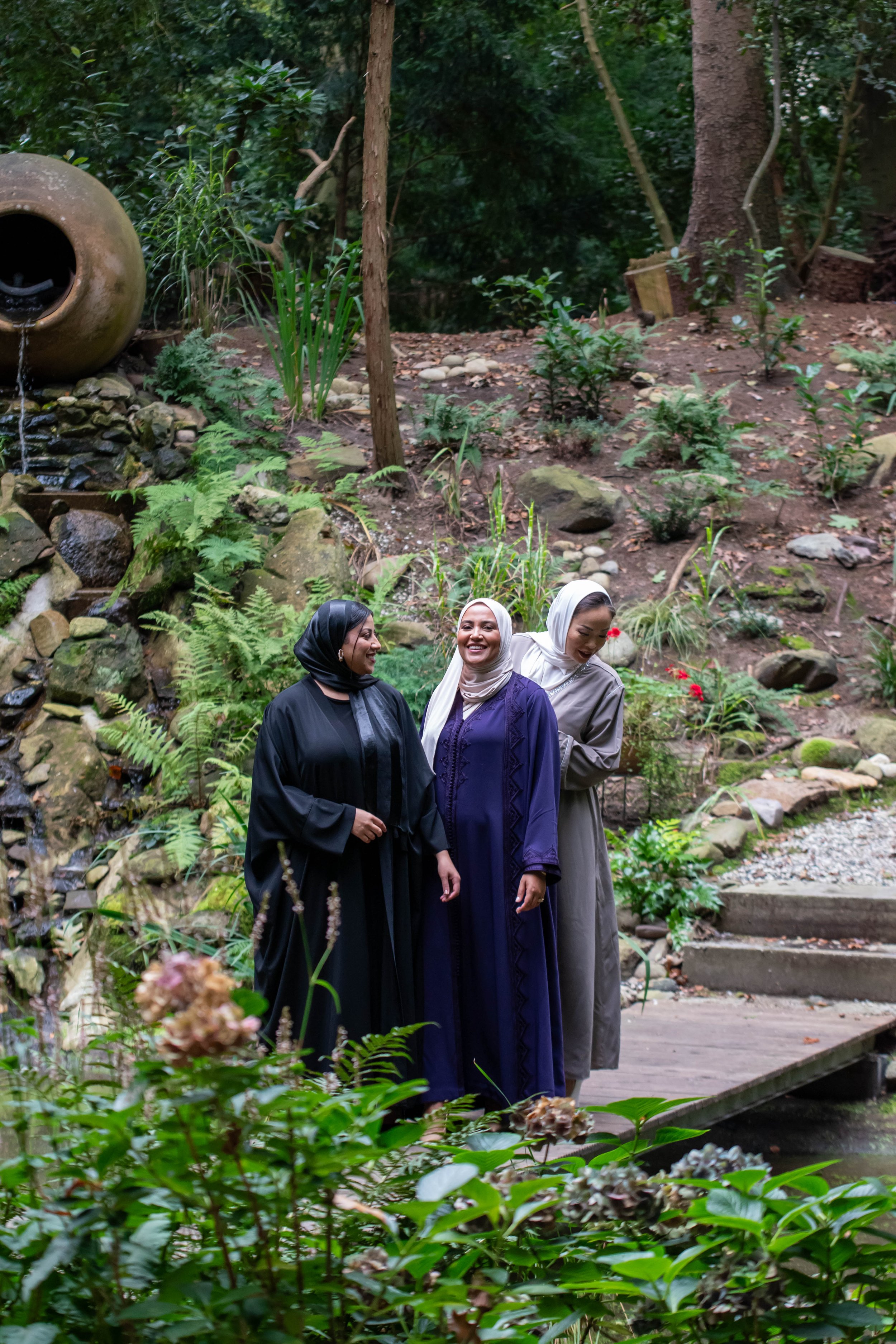Three women in modest clothes and headscarves laugh and talk on a wooden bridge in a garden with green plants, trees and rocks.