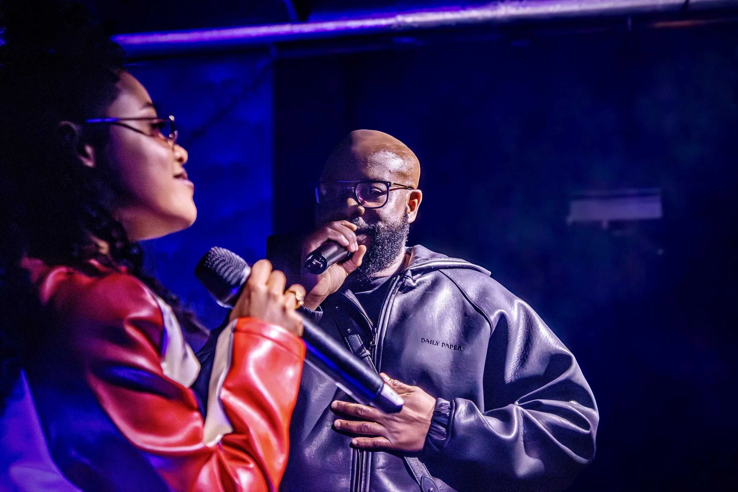 A woman and a man are on stage, both holding microphones. The woman has dark hair and is wearing glasses and a red and white jacket. The man has a beard, glasses, and is wearing a black leather jacket. The background is dark with blue lighting.