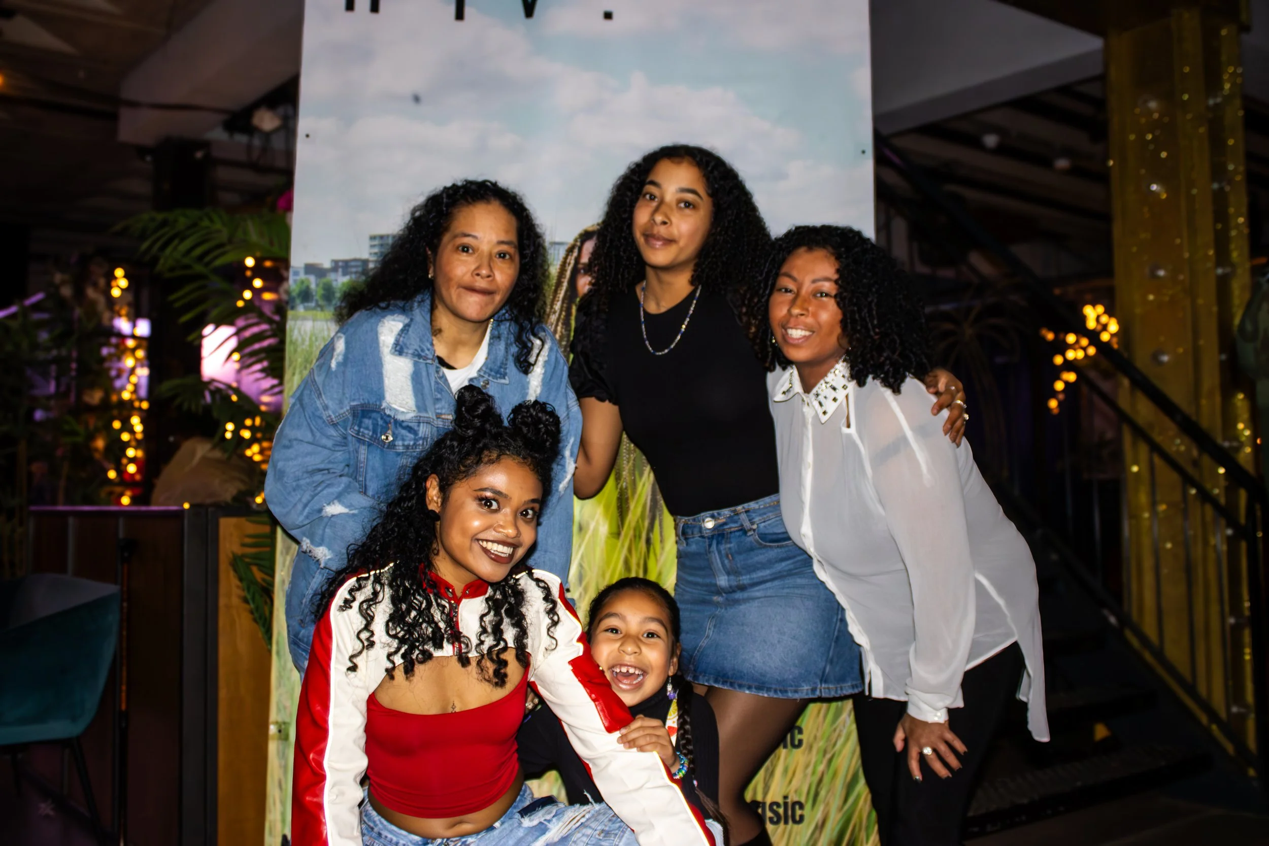 Group of six women and one young girl smiling and posing together indoors near a backdrop of a cloudy sky and cityscape, decorated with string lights and plants.