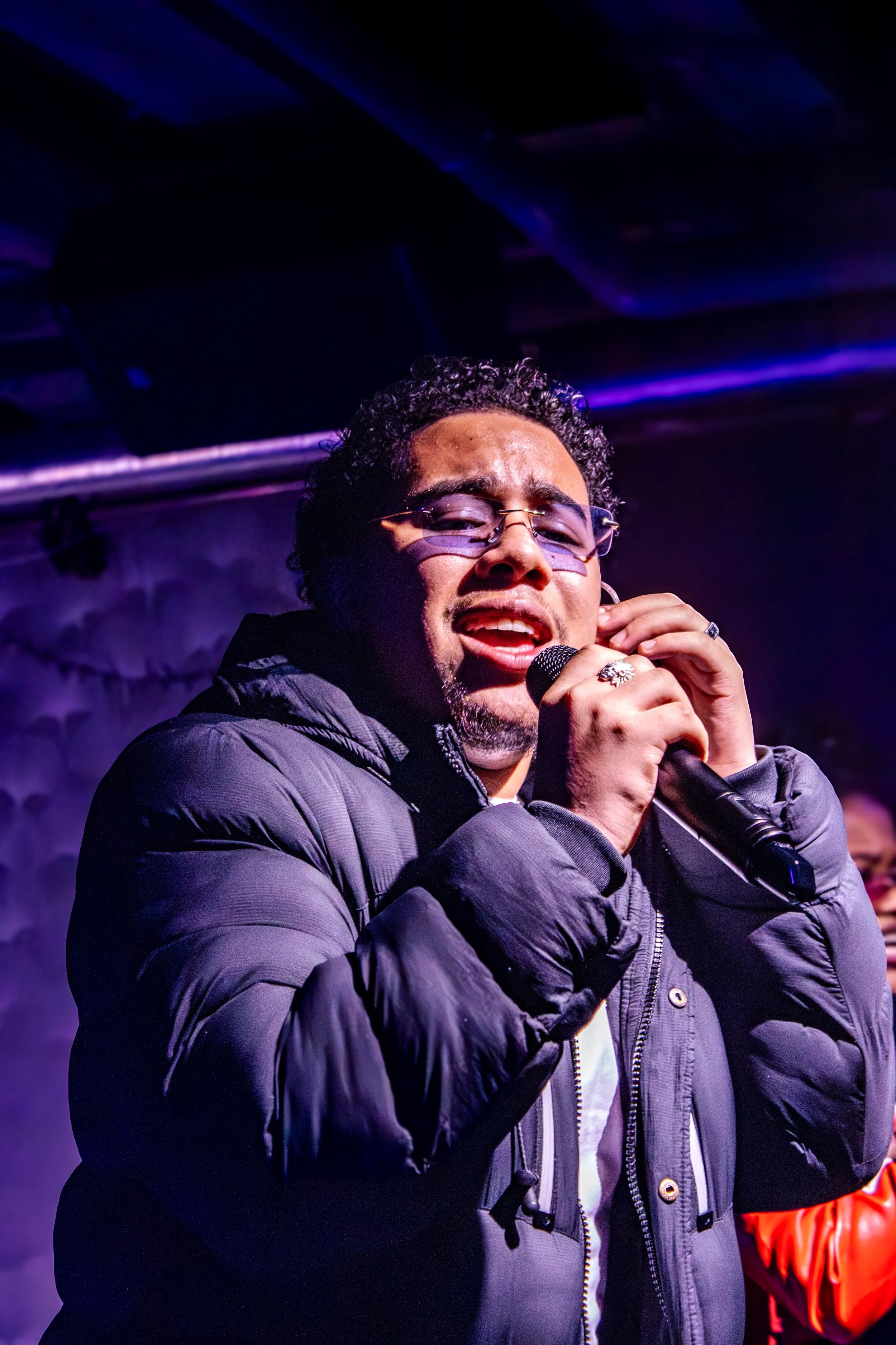 A man singing into a microphone on stage, wearing glasses and a black puffer jacket, with purple lighting in the background.