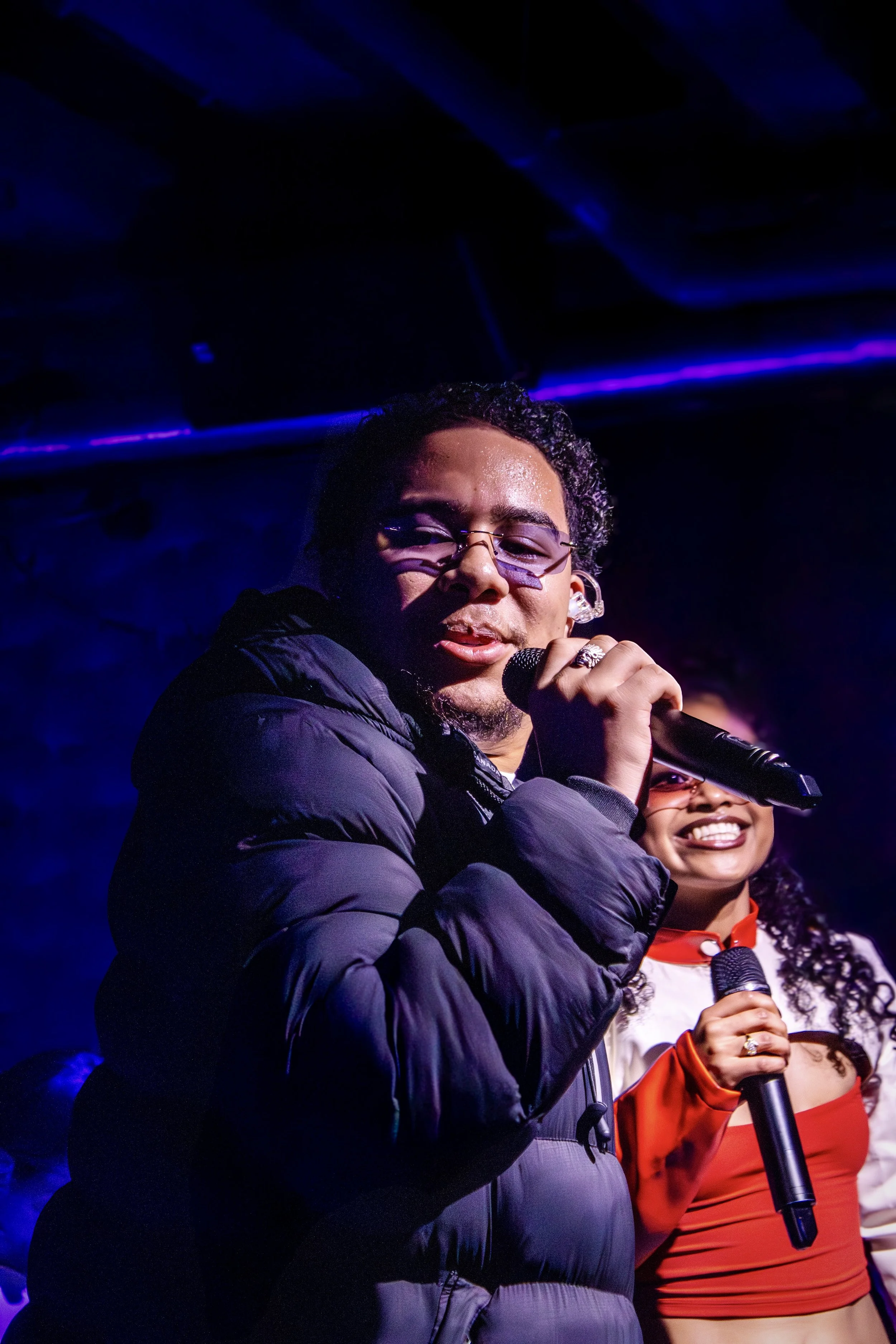 Man and woman performing on stage, both holding microphones, with the man wearing glasses and a puffy jacket, and the woman smiling in a red and white outfit.