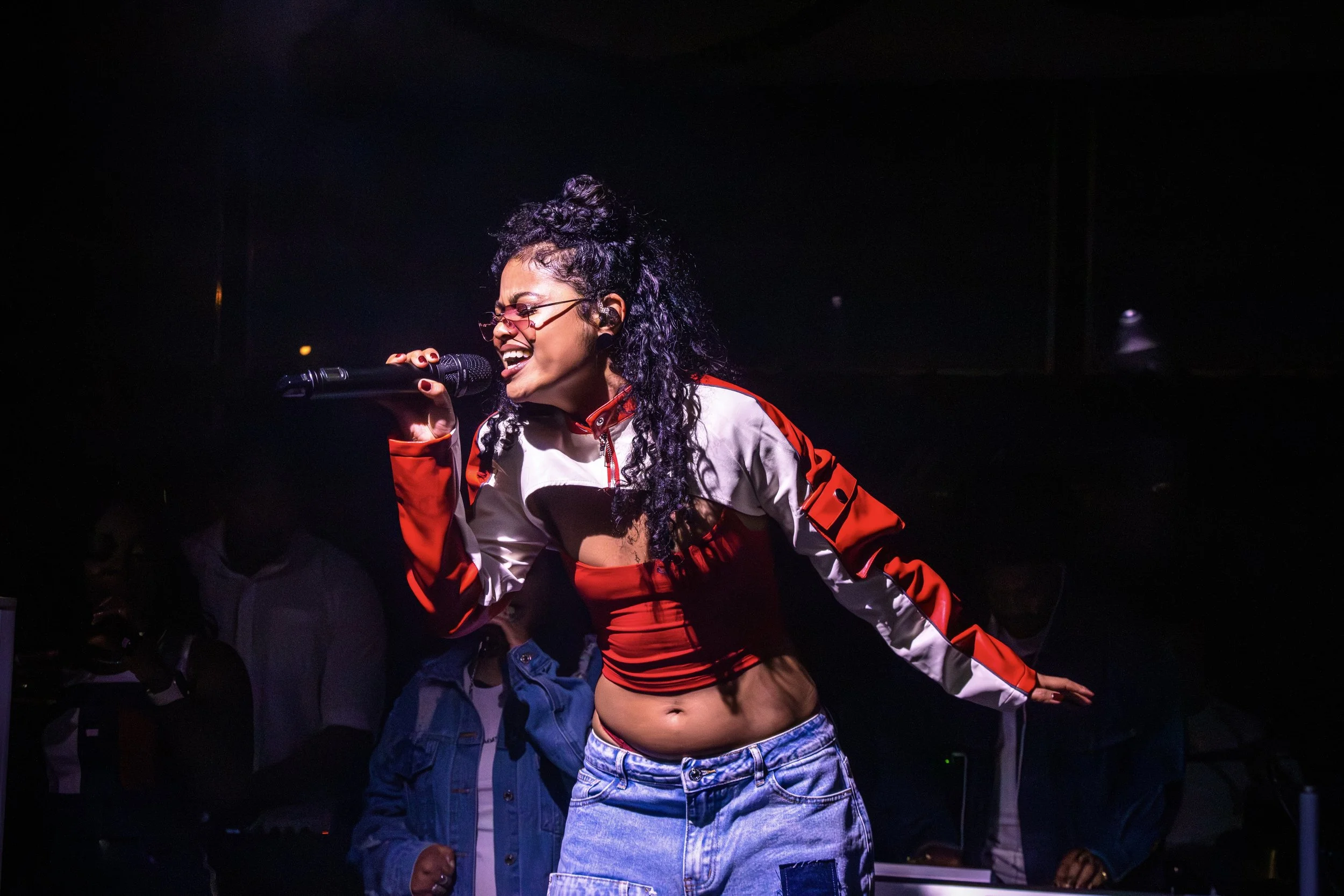 A woman passionately singing into a microphone on stage, wearing a red and white jacket and baggy jeans, with glasses and curly hair, in a dimly lit environment.