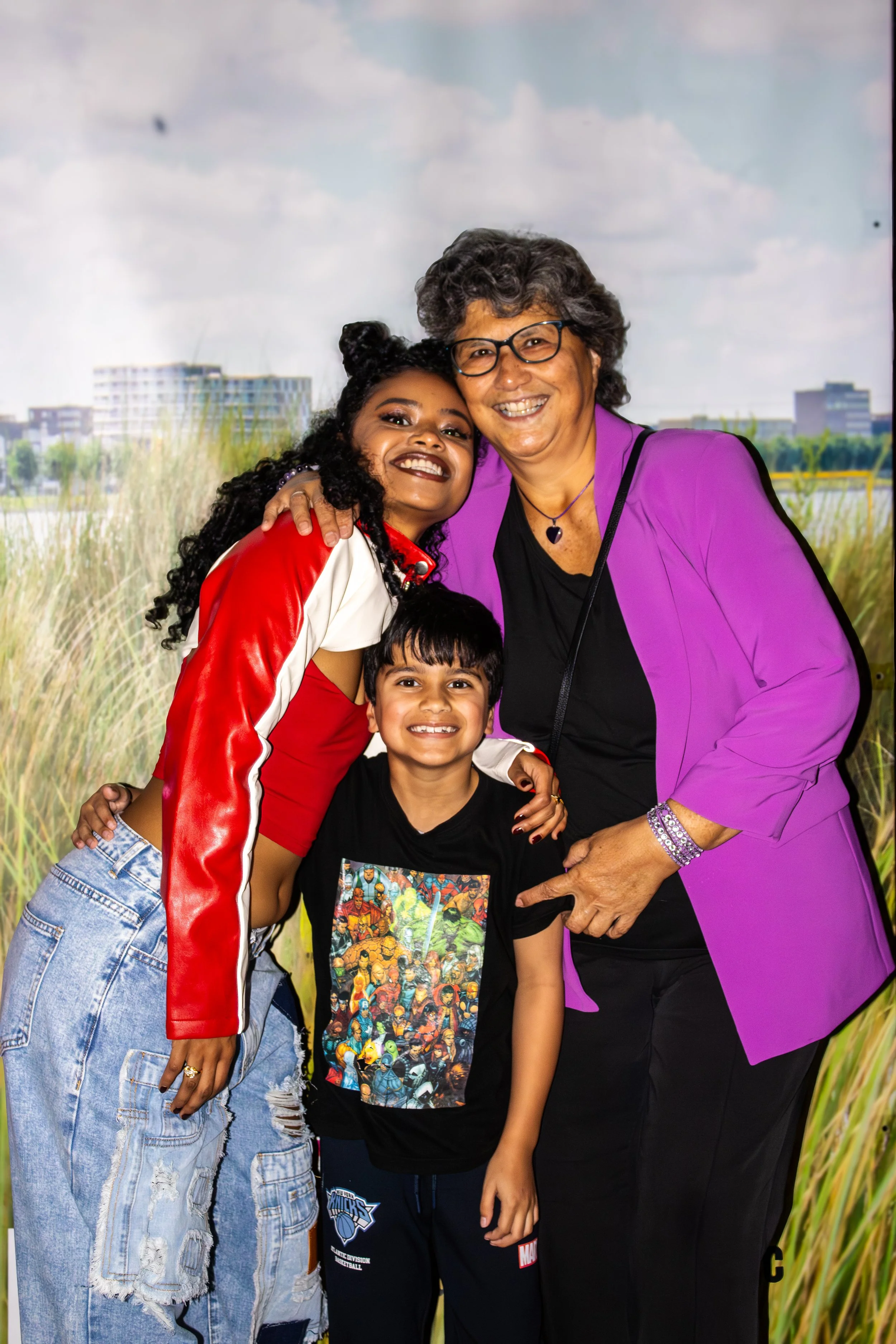 A happy multigenerational family portrait featuring two women and two kids, smiling and hugging in front of a natural outdoor backdrop with tall grasses and buildings in the distance.