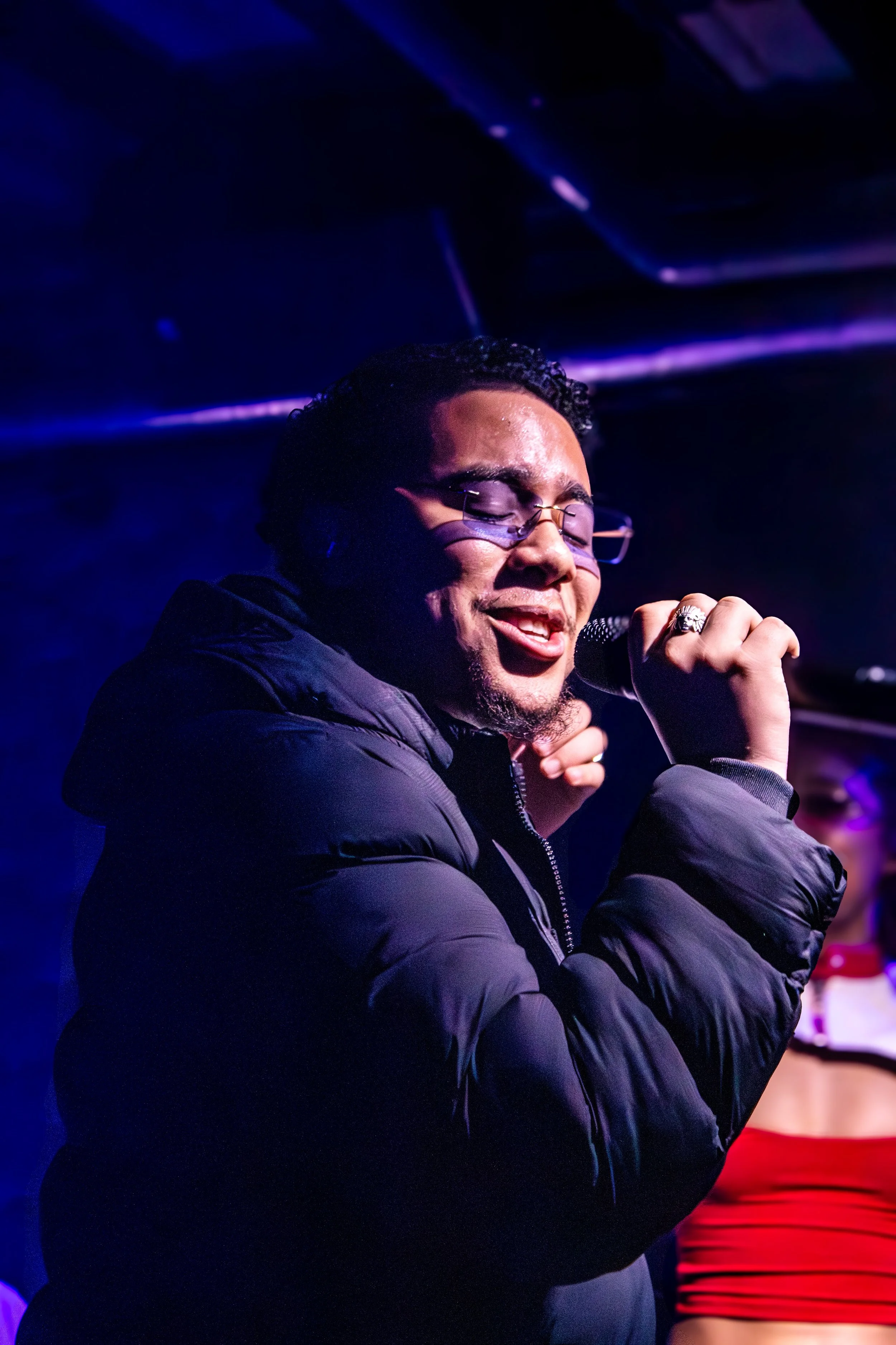 Man singing into microphone with eyes closed, wearing glasses and a black puffy jacket, on stage with purple and blue lighting.