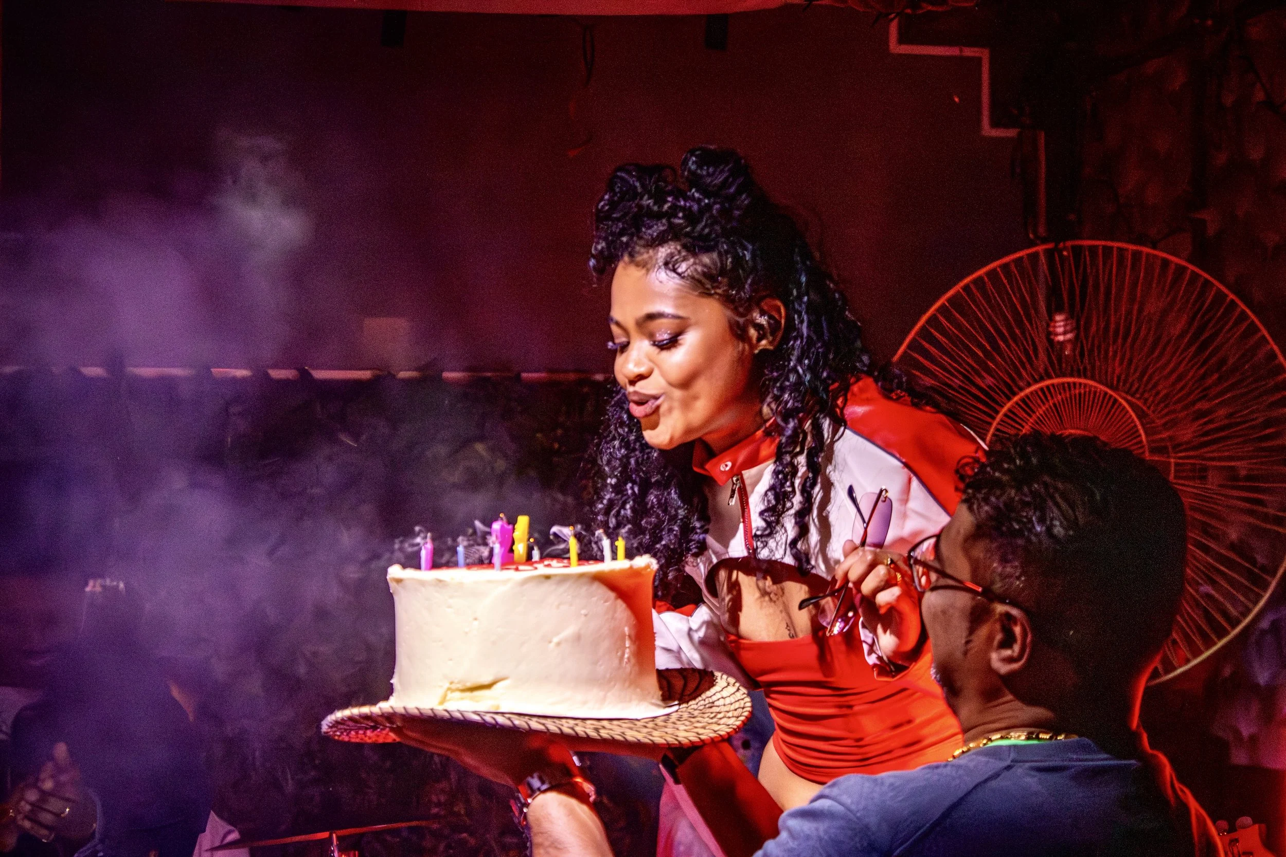 Woman with curly hair celebrating her birthday with a cake, standing next to a man wearing glasses, in a dimly lit room with purple and red lighting.