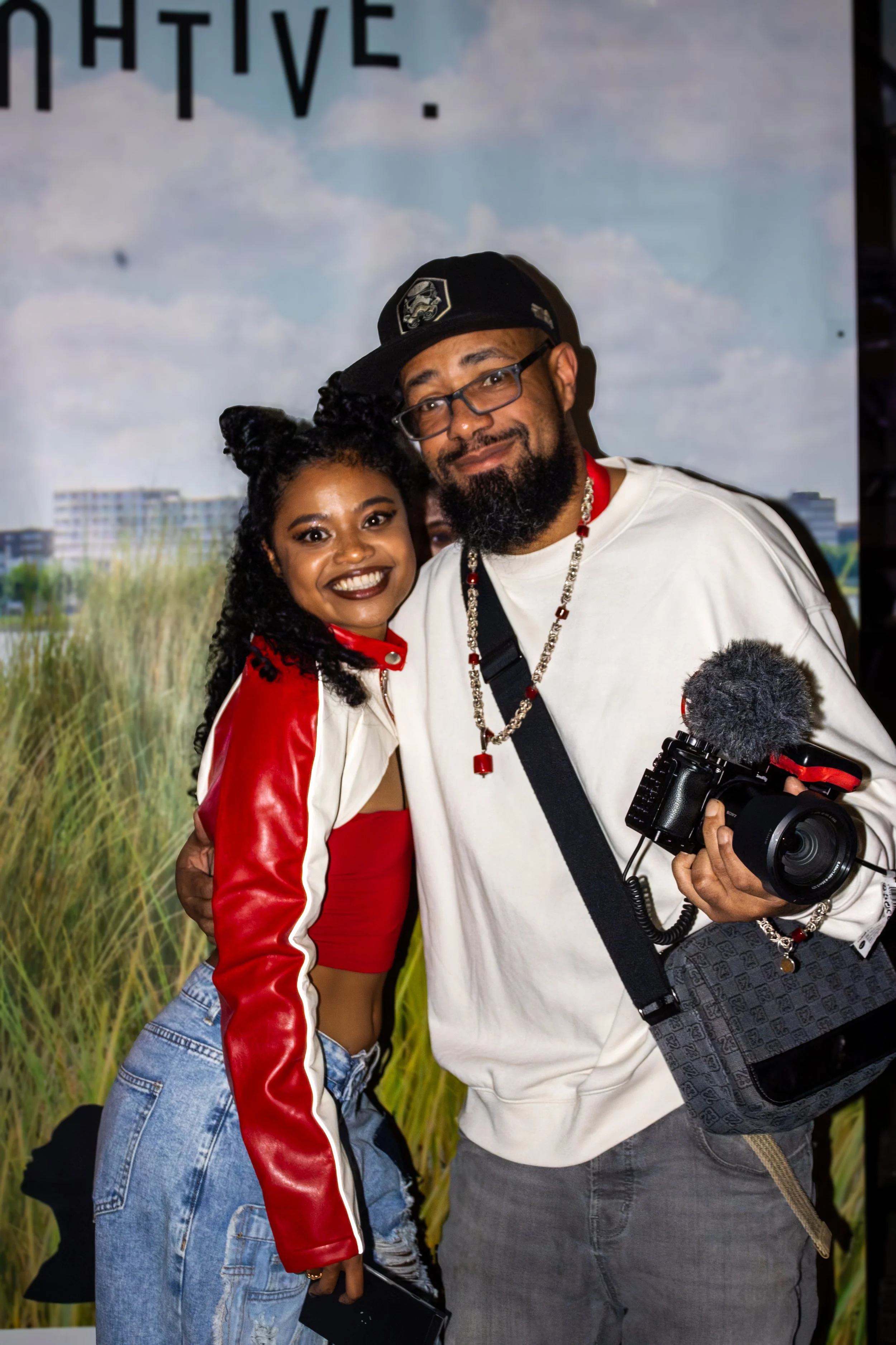 A young woman with dark hair styled in buns and wearing a red jacket and jeans poses with a bearded man with glasses, wearing a white sweatshirt, cap, and a camera. They are smiling and standing in front of a backdrop with a nature scene and building
