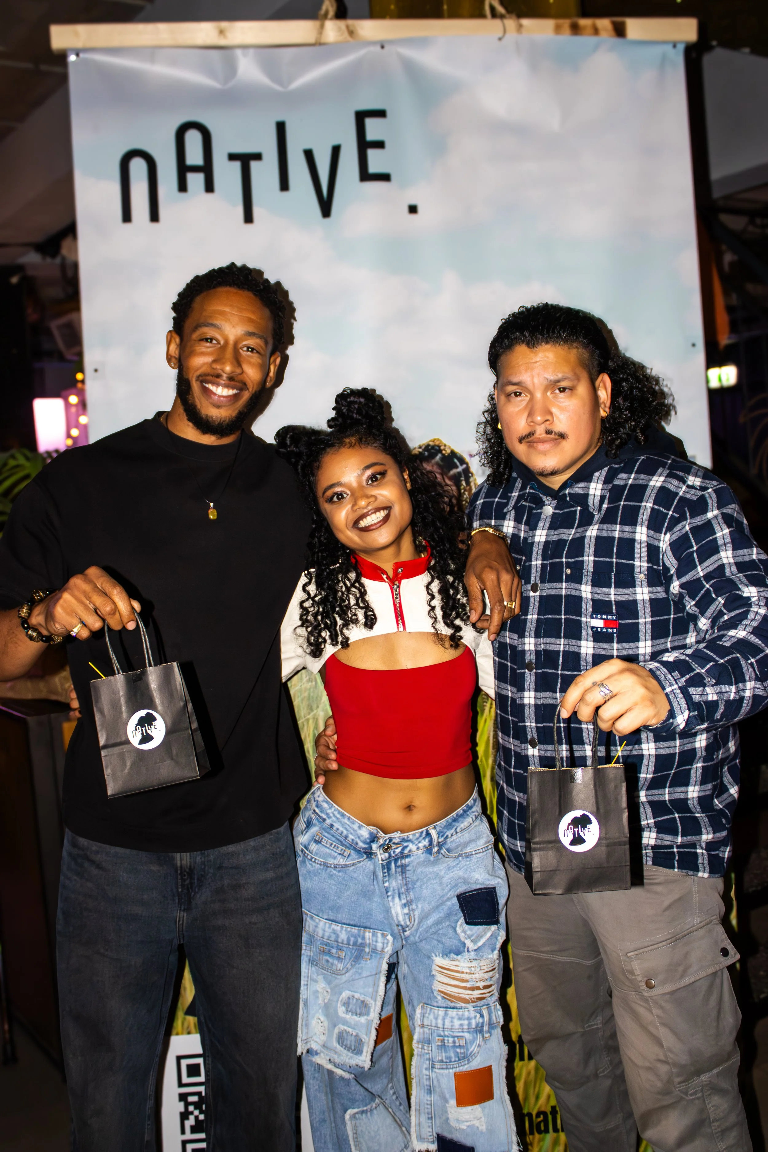 Three people standing side by side holding black bags with a white logo, smiling for the camera in front of a backdrop that says 'Native' with a blue sky and clouds.