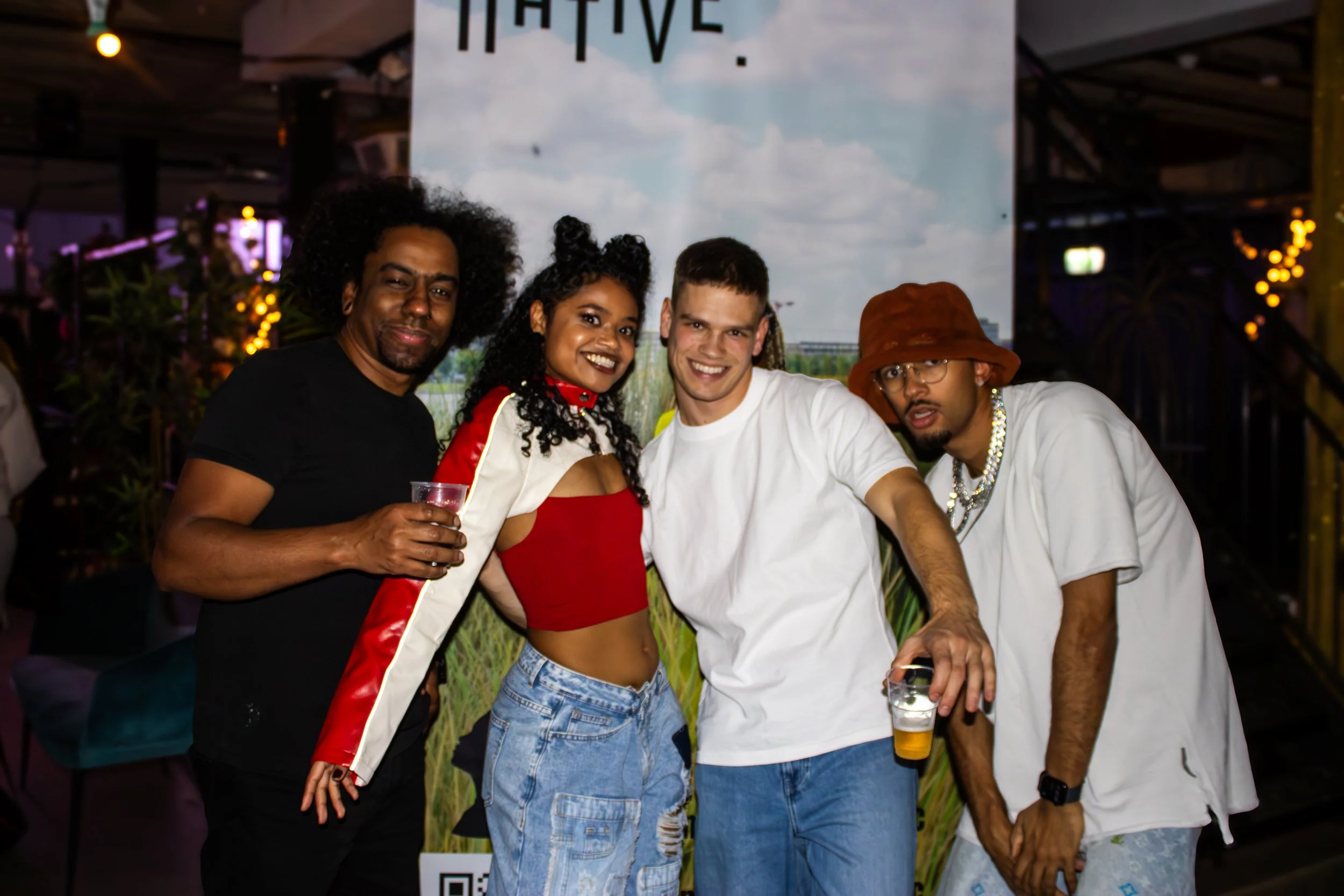 Four people standing together in a social setting, smiling, with drinks in their hands. The background has a decorative banner and warm lighting.