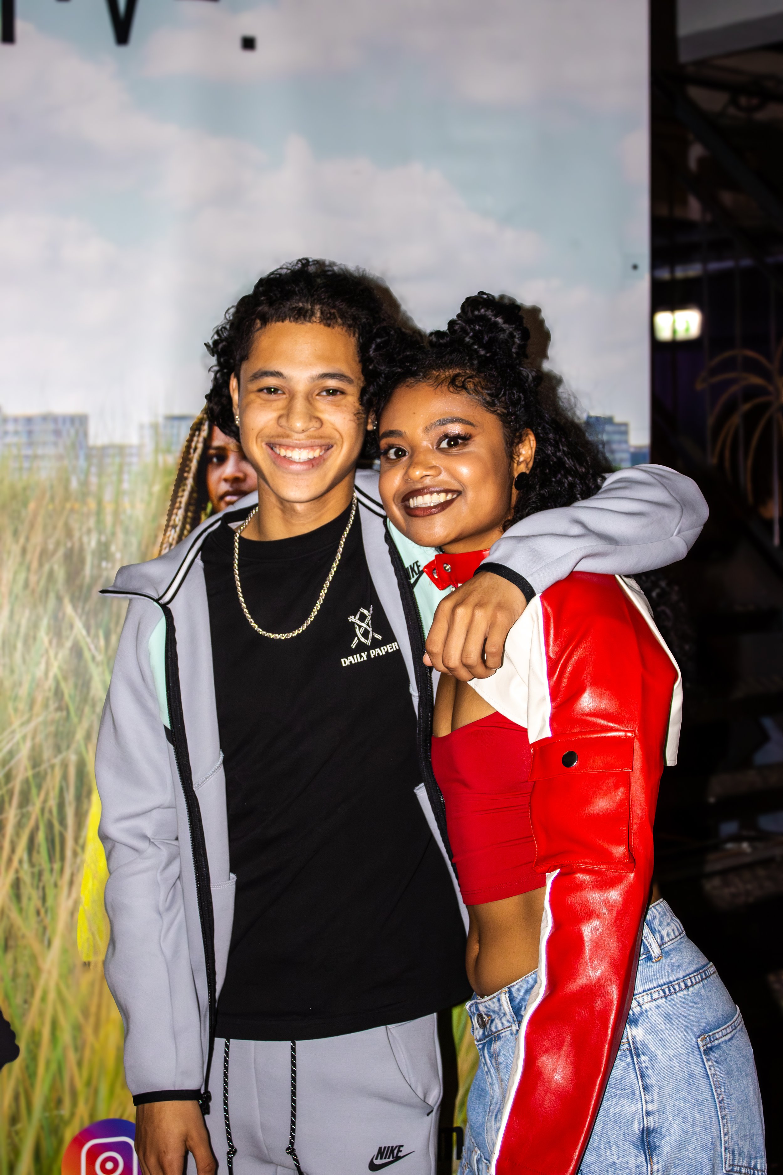 Two young adults smiling and posing for a photo, one with an arm around the other's shoulder. They are standing in front of a backdrop with a landscape scene.