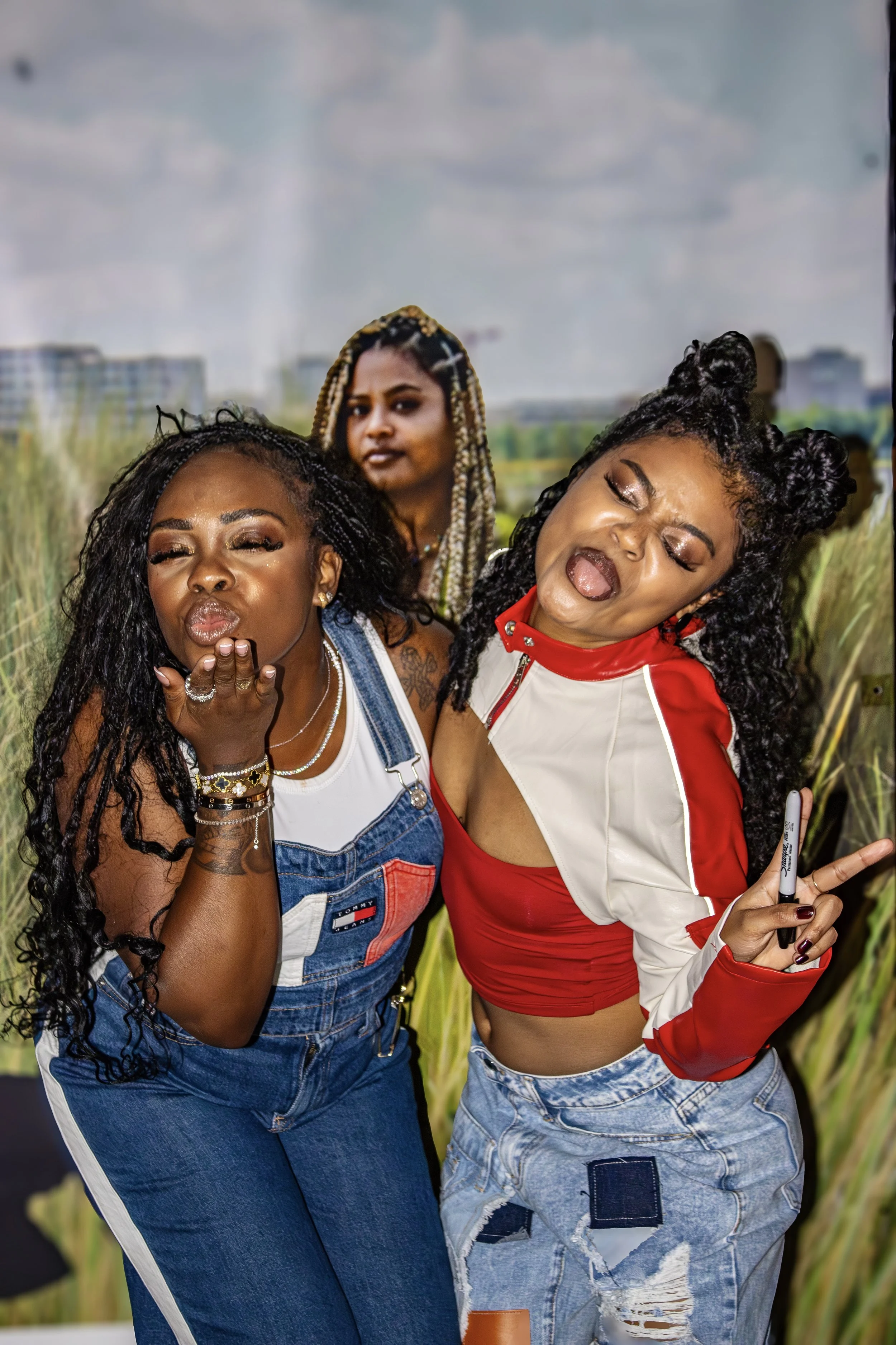 Three women posing playfully outdoors near tall grass and a cityscape in the background.