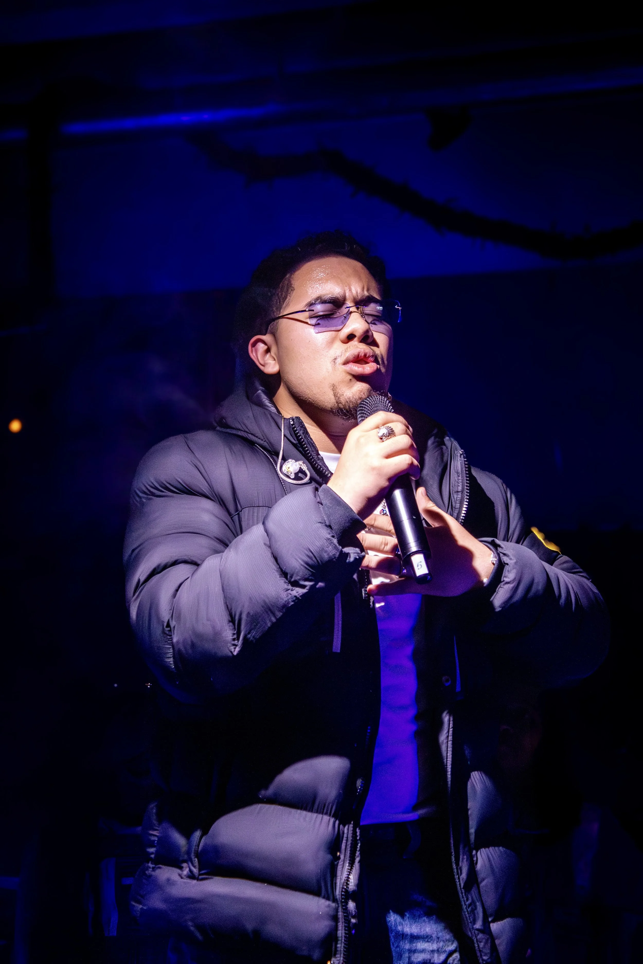 A young man wearing glasses, a puffy jacket, singing or speaking into a microphone with his hand on his chest, under blue and purple lighting.