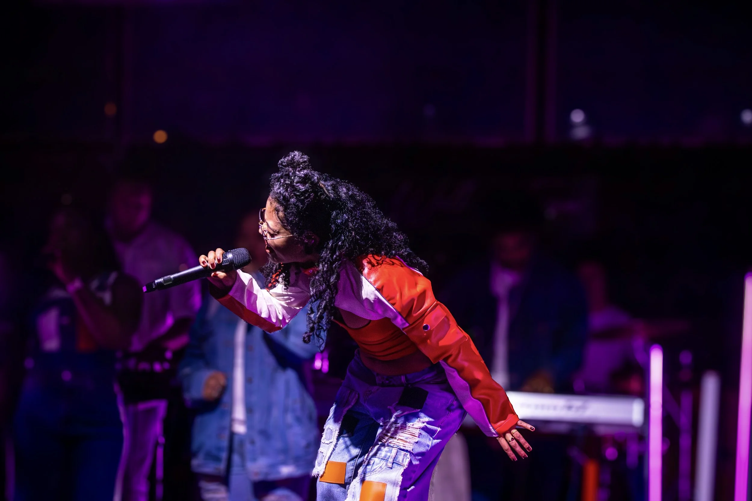 A woman with curly black hair and glasses singing into a microphone on stage, wearing a red and white jacket and distressed jeans, with a purple and pink lit background and several people in the background.