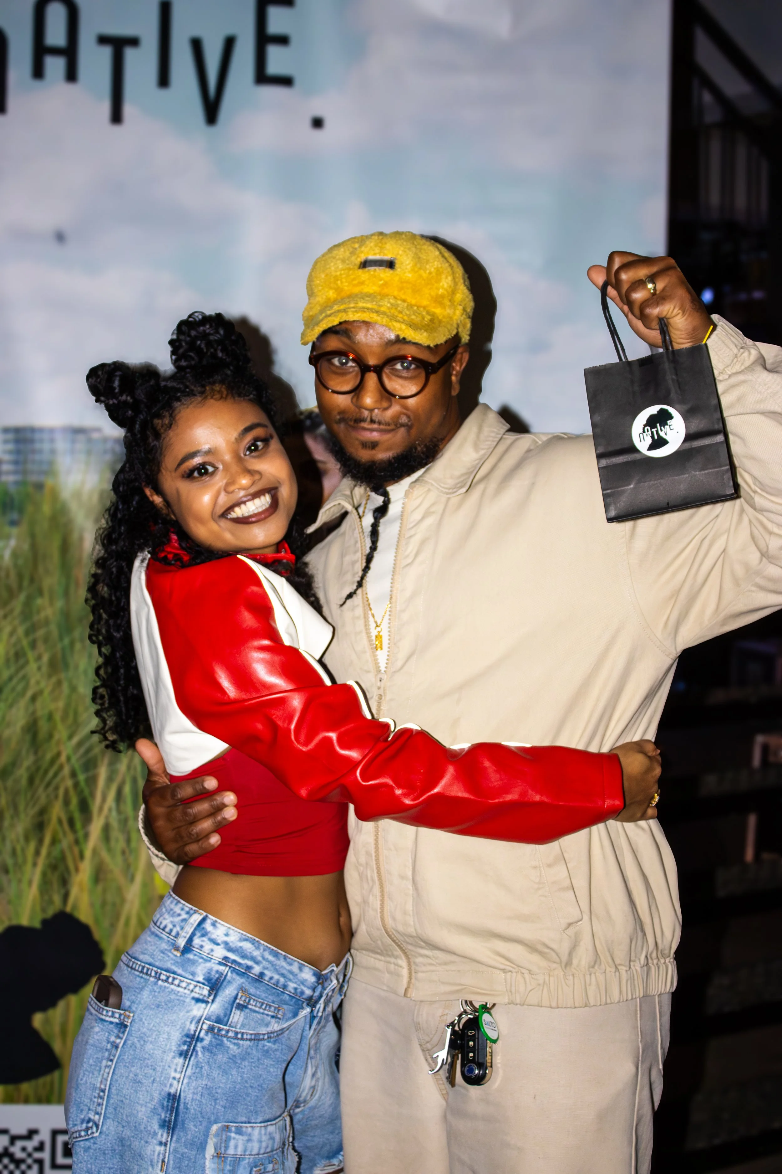 A happy couple hugging, with the woman wearing a red and white cropped jacket and jeans, and the man holding a small black gift bag.