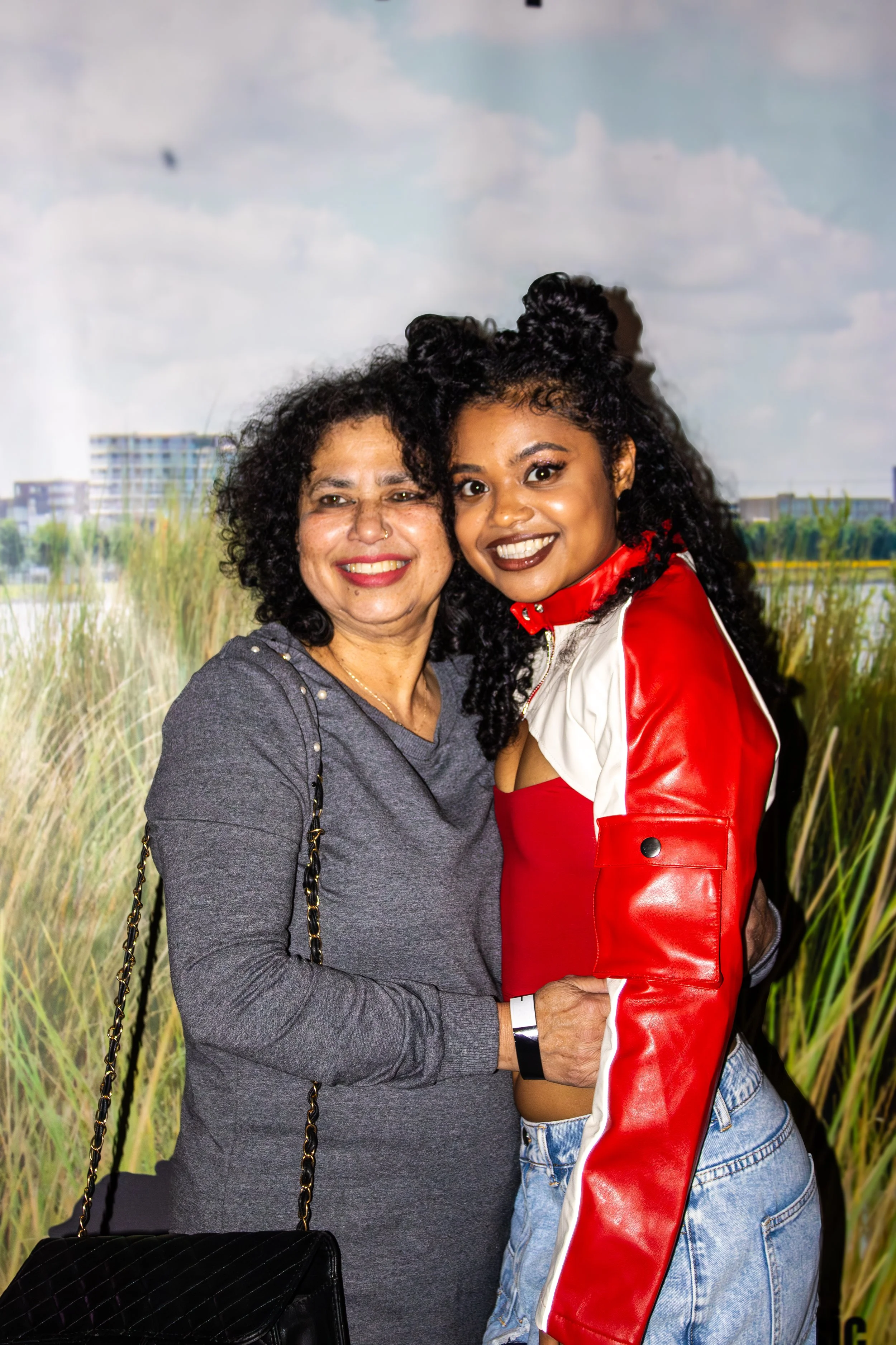 Two women smiling and hugging against a backdrop of a grassy field and a cloudy sky.