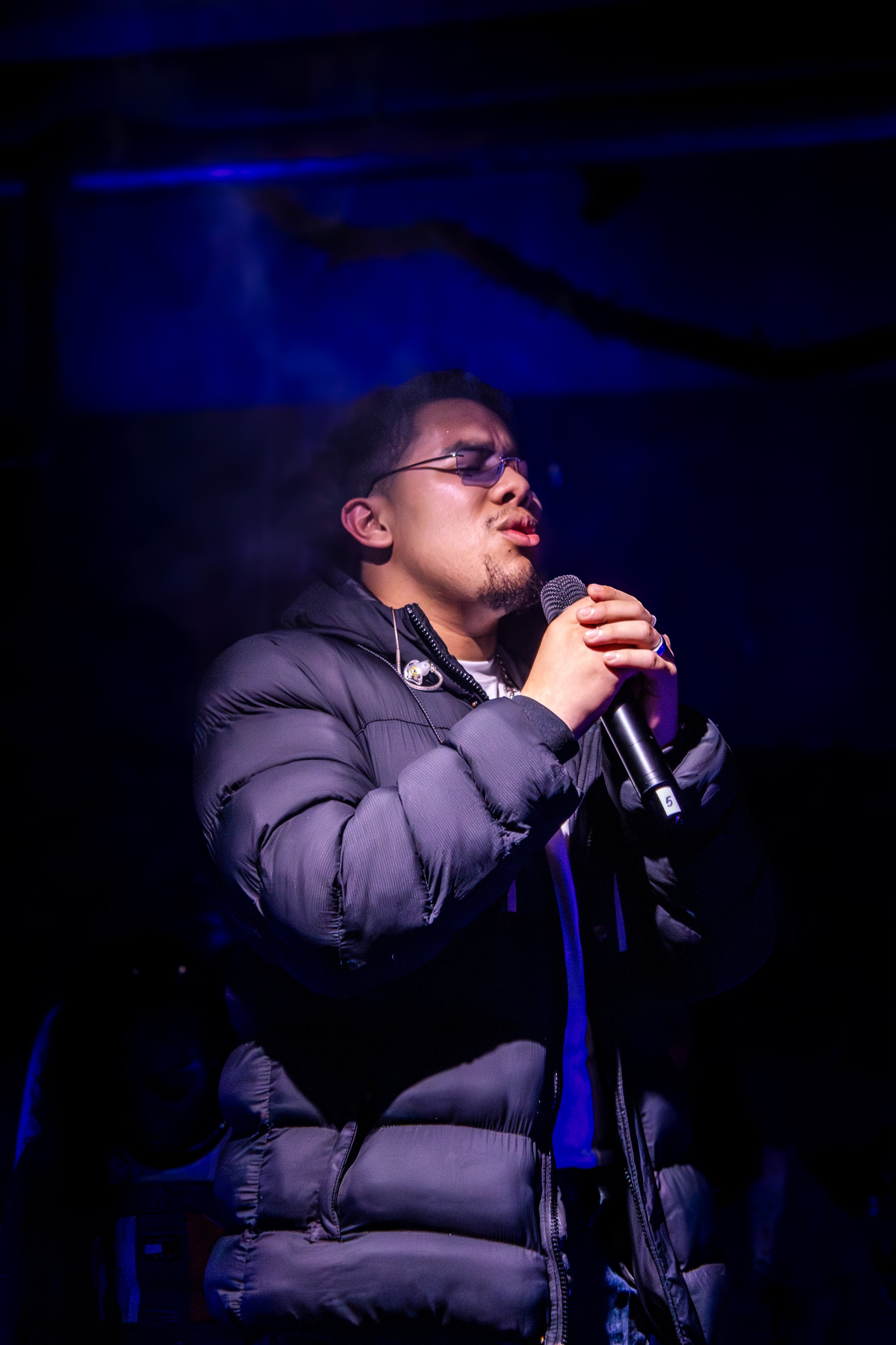 A man singing into a microphone during a performance, wearing glasses and a puffy jacket, with dark background lighting.