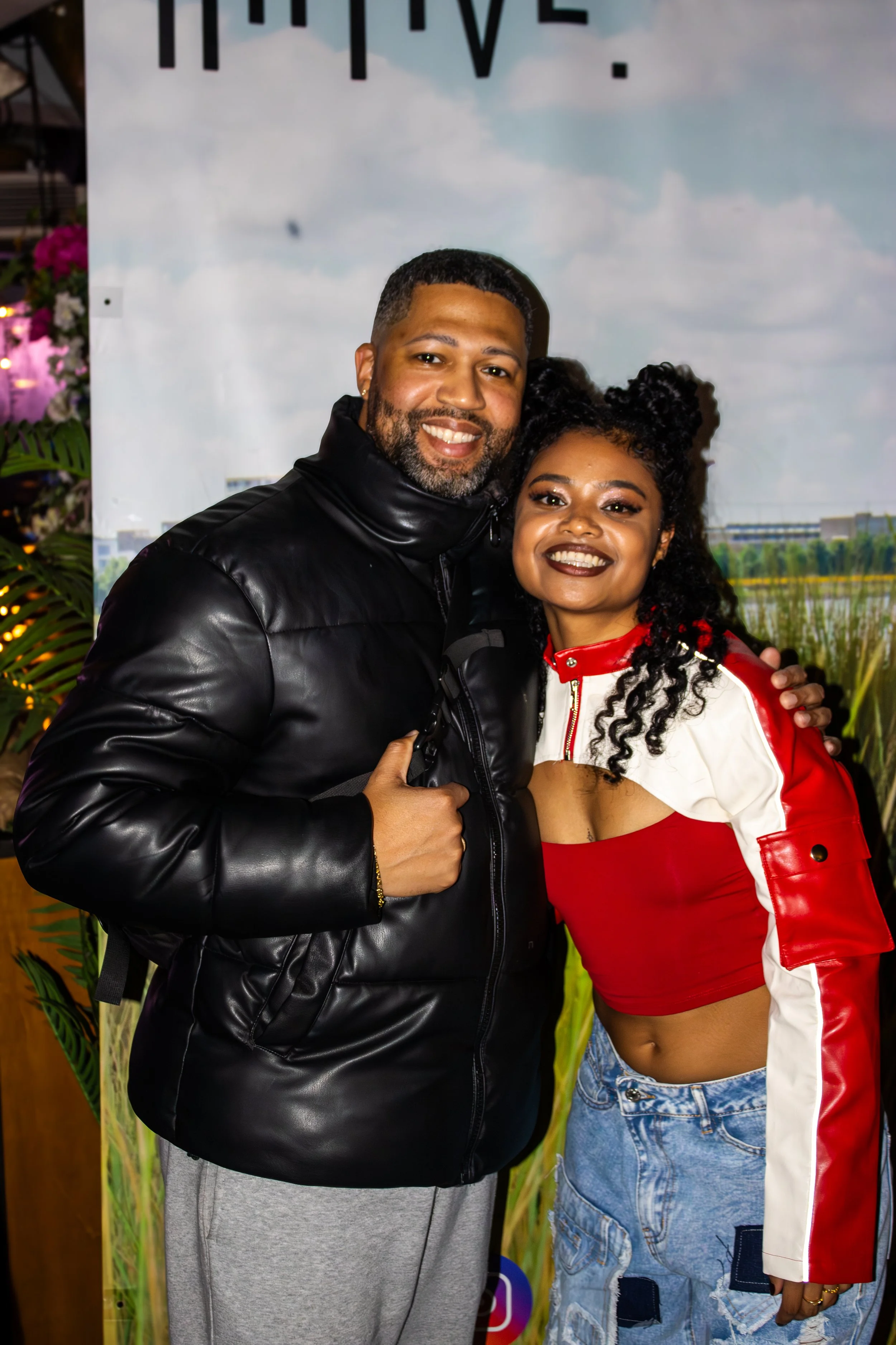 A smiling Black man and woman posing together in front of a scenic backdrop. The man is wearing a black leather jacket and grey pants, giving a thumbs up. The woman is dressed in a red crop top with white and red sleeves and distressed jeans, with he