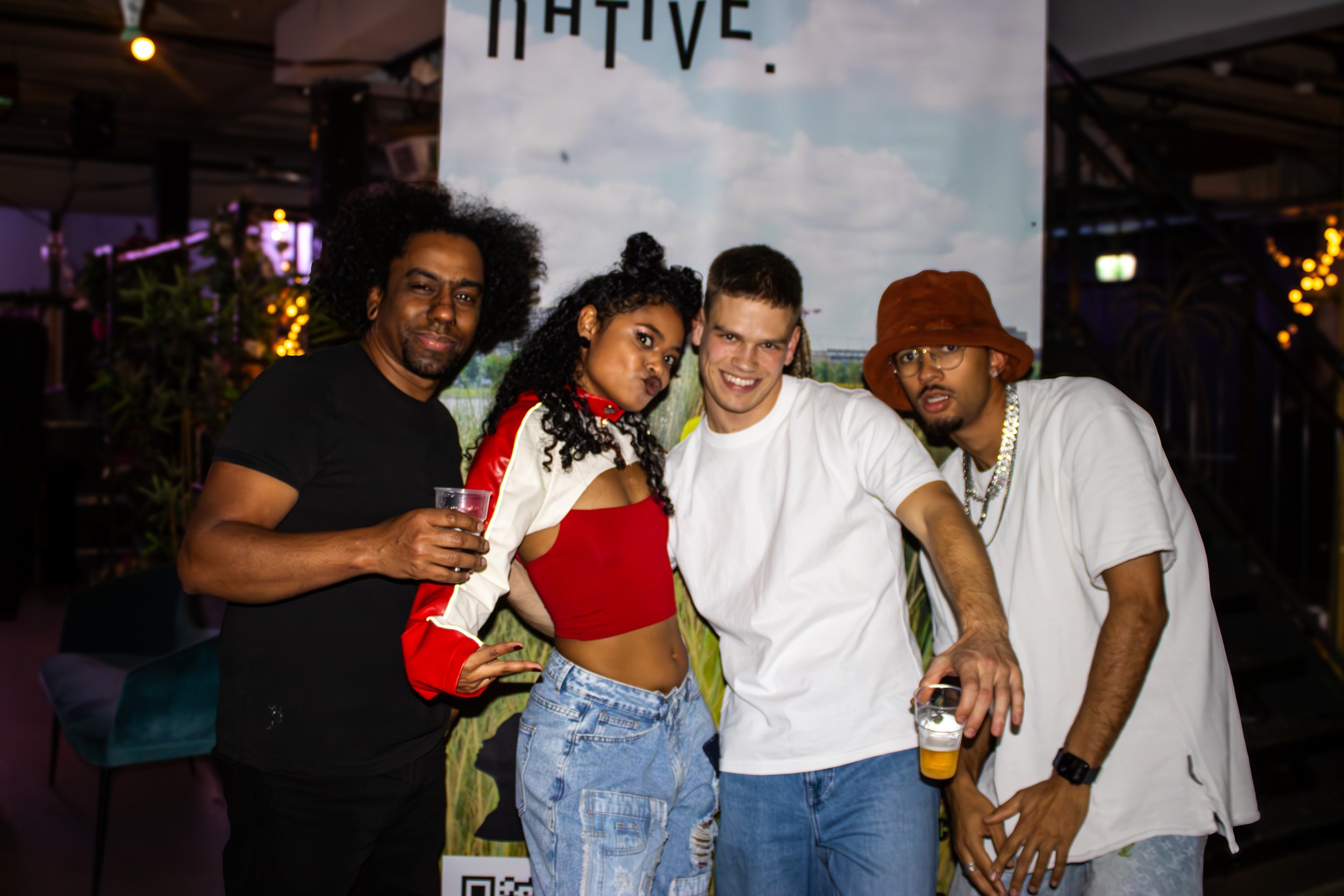 Four people standing together at a social event, holding drinks, smiling, and posing for the camera with a large banner behind them that says 'NATIVE' and has a sky and clouds background.