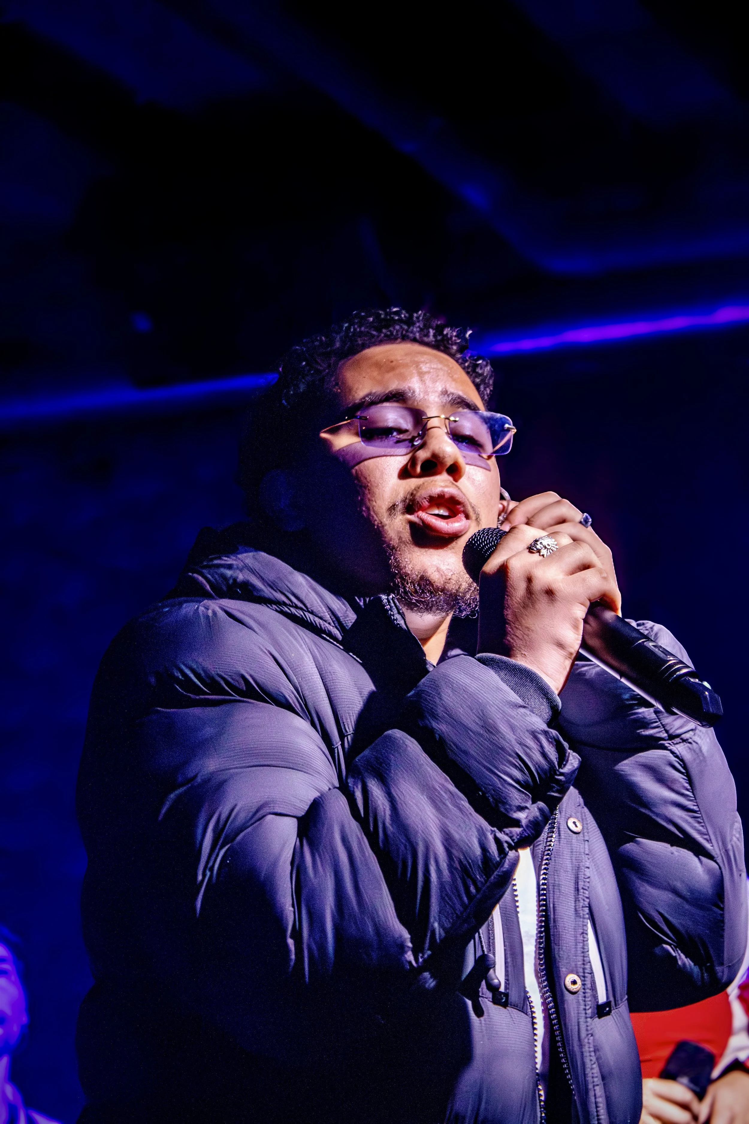 A person singing into a microphone on stage, wearing glasses and a puffy jacket, with dark lighting and purple hues in the background.