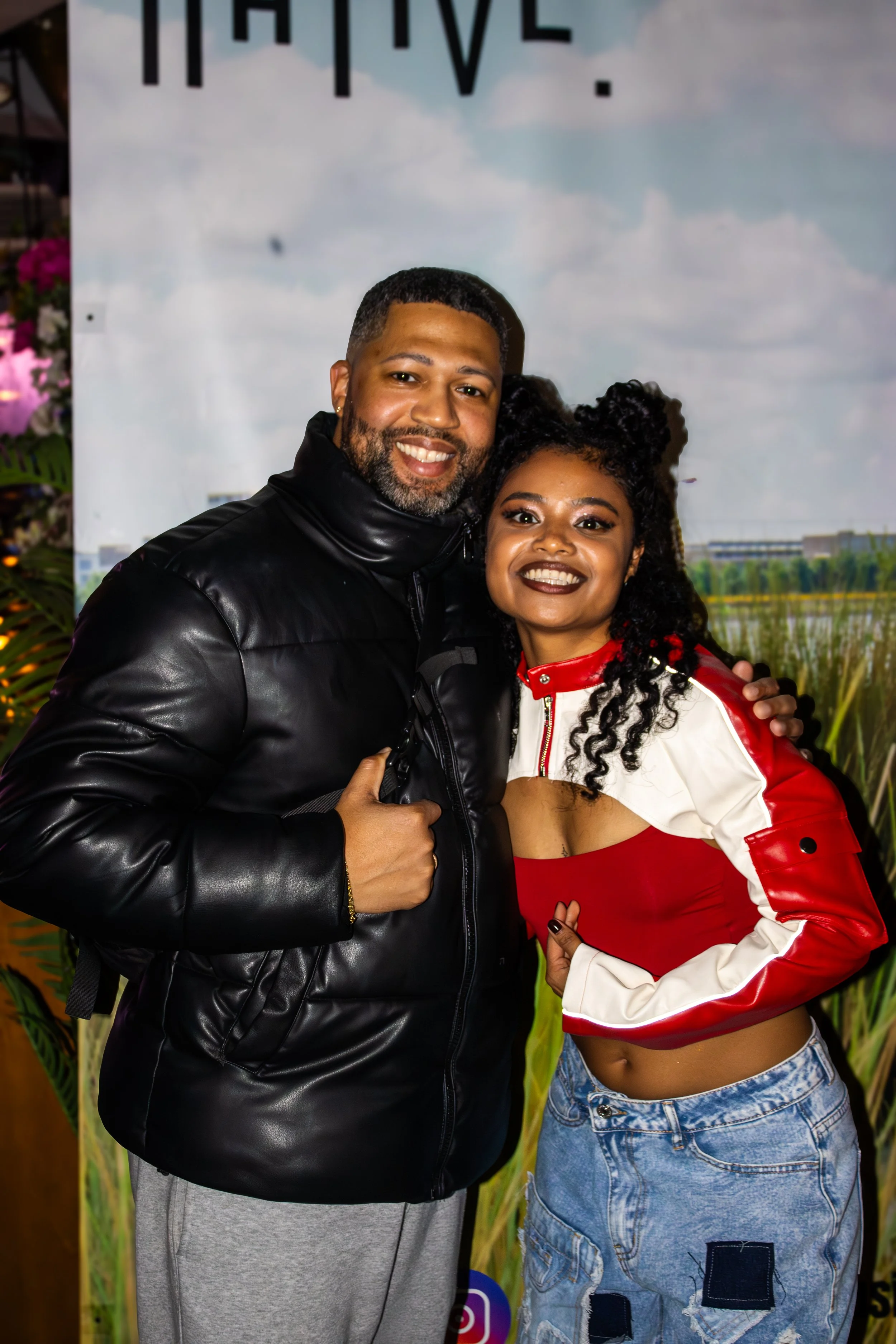 A smiling couple posing together, the man wearing a black leather jacket and giving a thumbs up, the woman in a red and white cropped jacket and distressed jeans, standing in front of a background with a lake and sky.