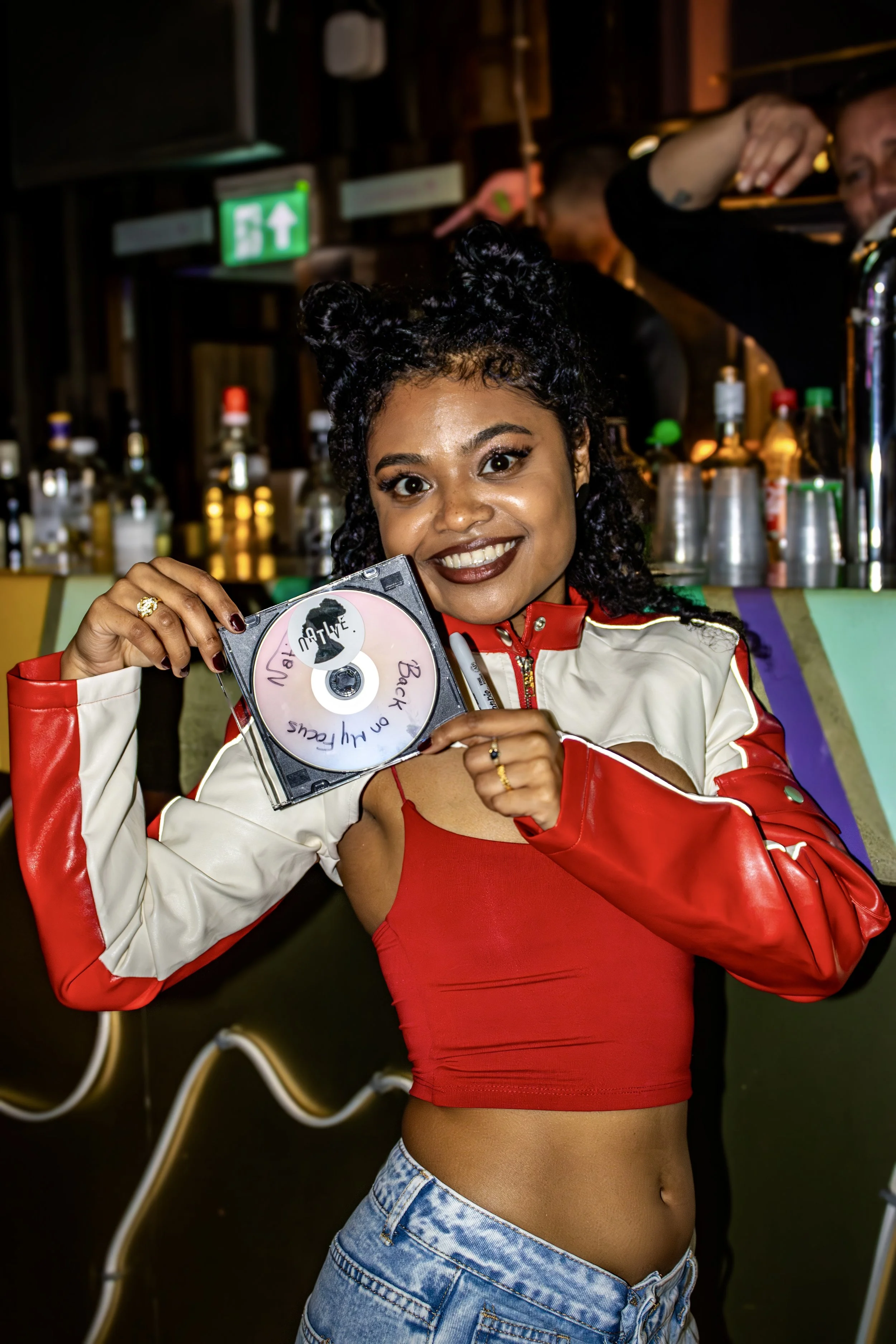 A young woman with curly hair styled in two buns is holding a CD with handwritten text that says, 'Back on the highway.' She is smiling and wearing a red tank top and a beige and red jacket, in a bar or club setting with various bottles of alcohol vi