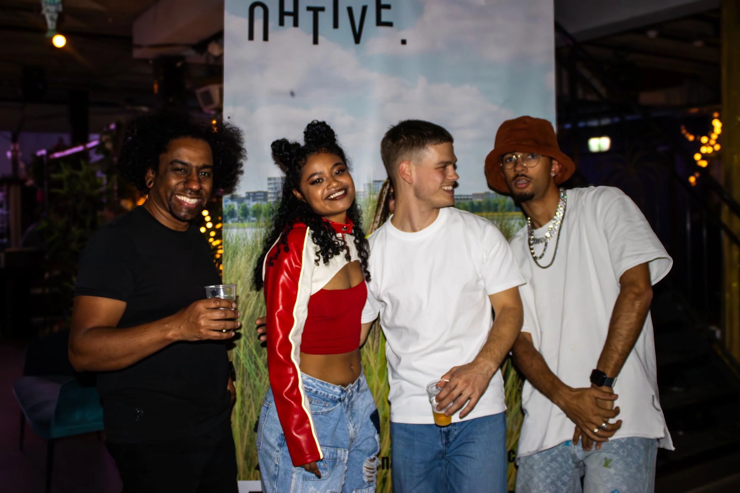 Four people at a social gathering smiling and holding drinks, standing outdoors in front of a banner with trees and a sky background.