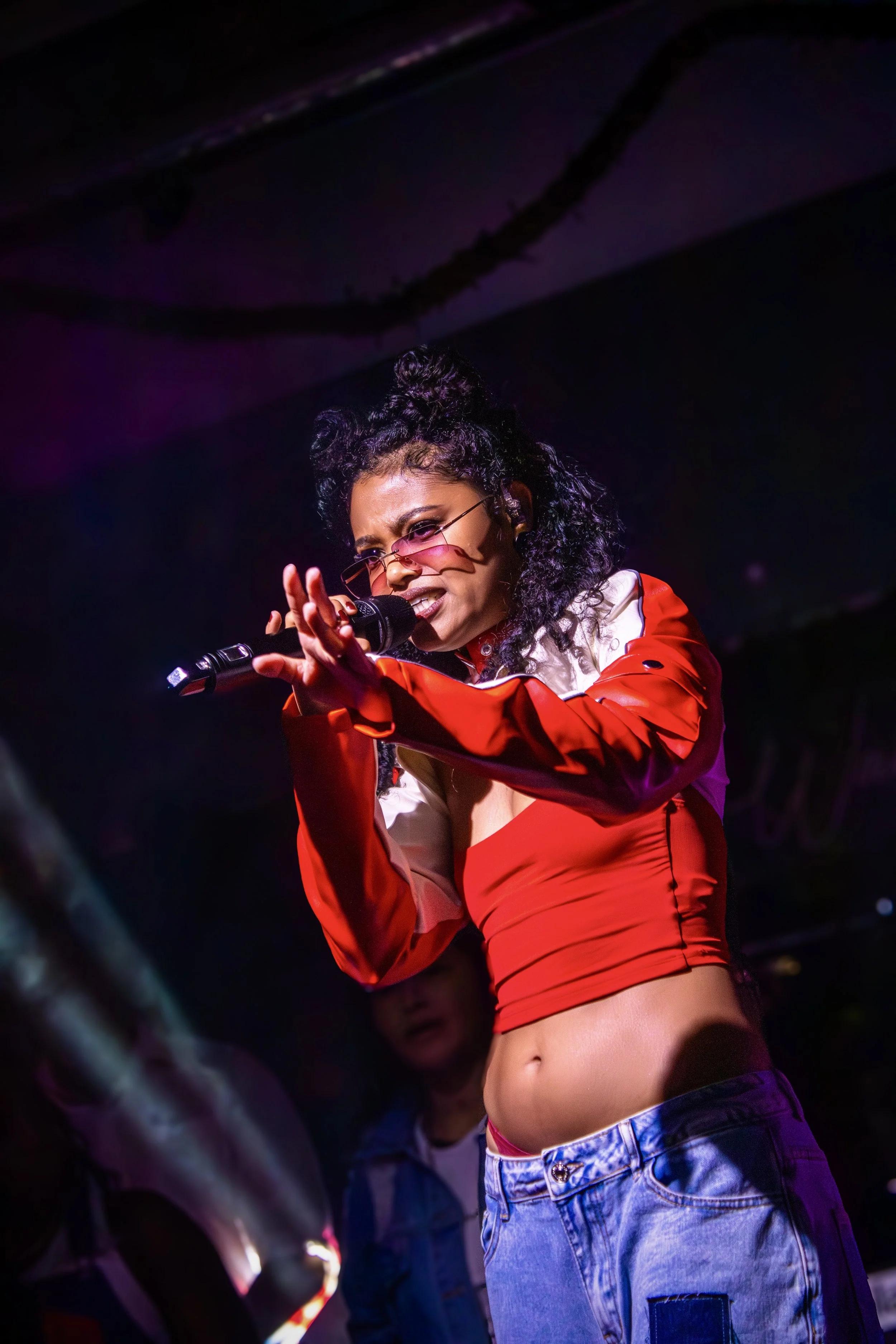 A woman singing into a microphone during a performance, wearing a red and white jacket, sunglasses, and jeans, with purple and pink stage lighting.