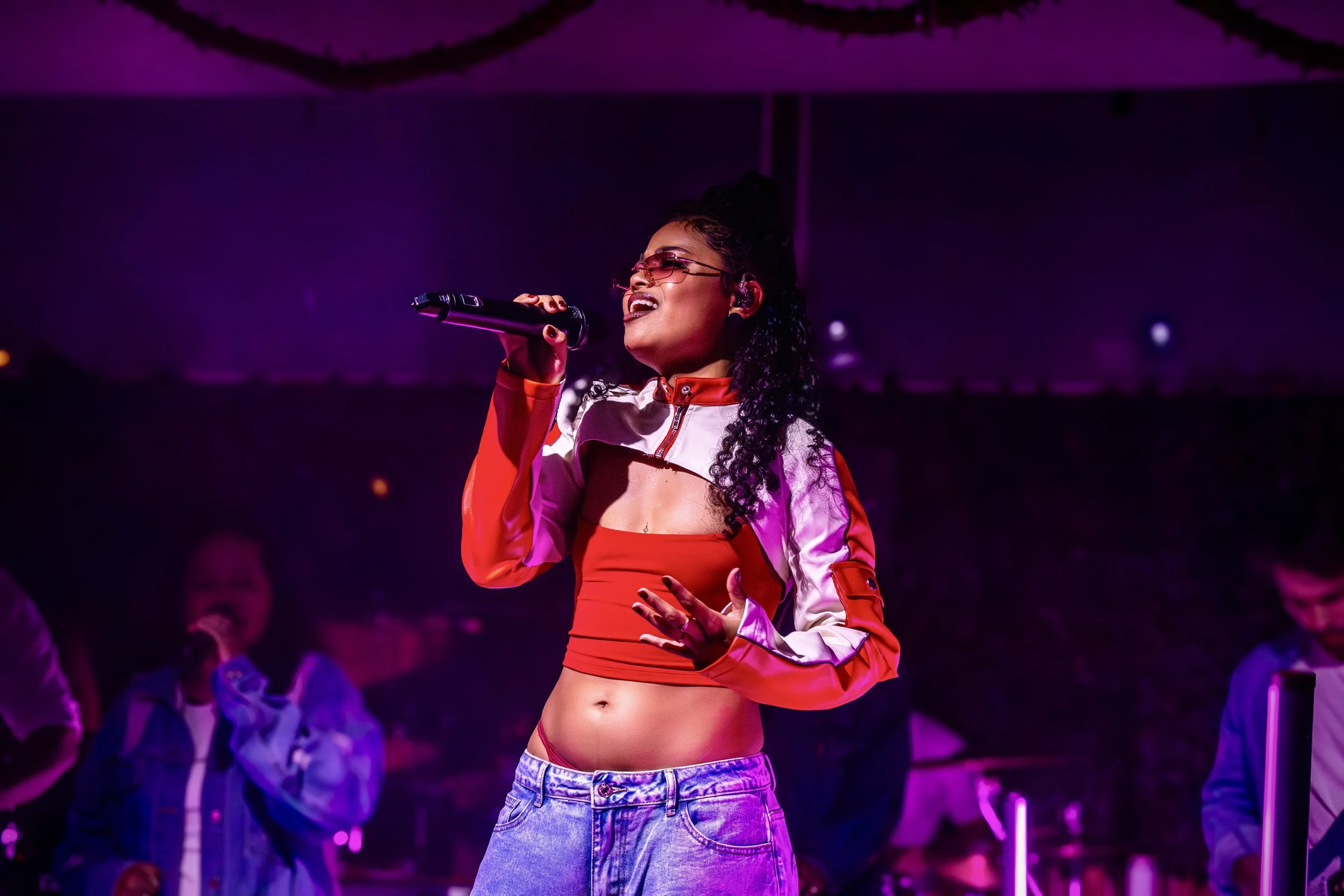 A woman singing on stage with purple lighting, wearing sunglasses, a red and white cropped jacket, a red crop top, and high-waisted jeans.