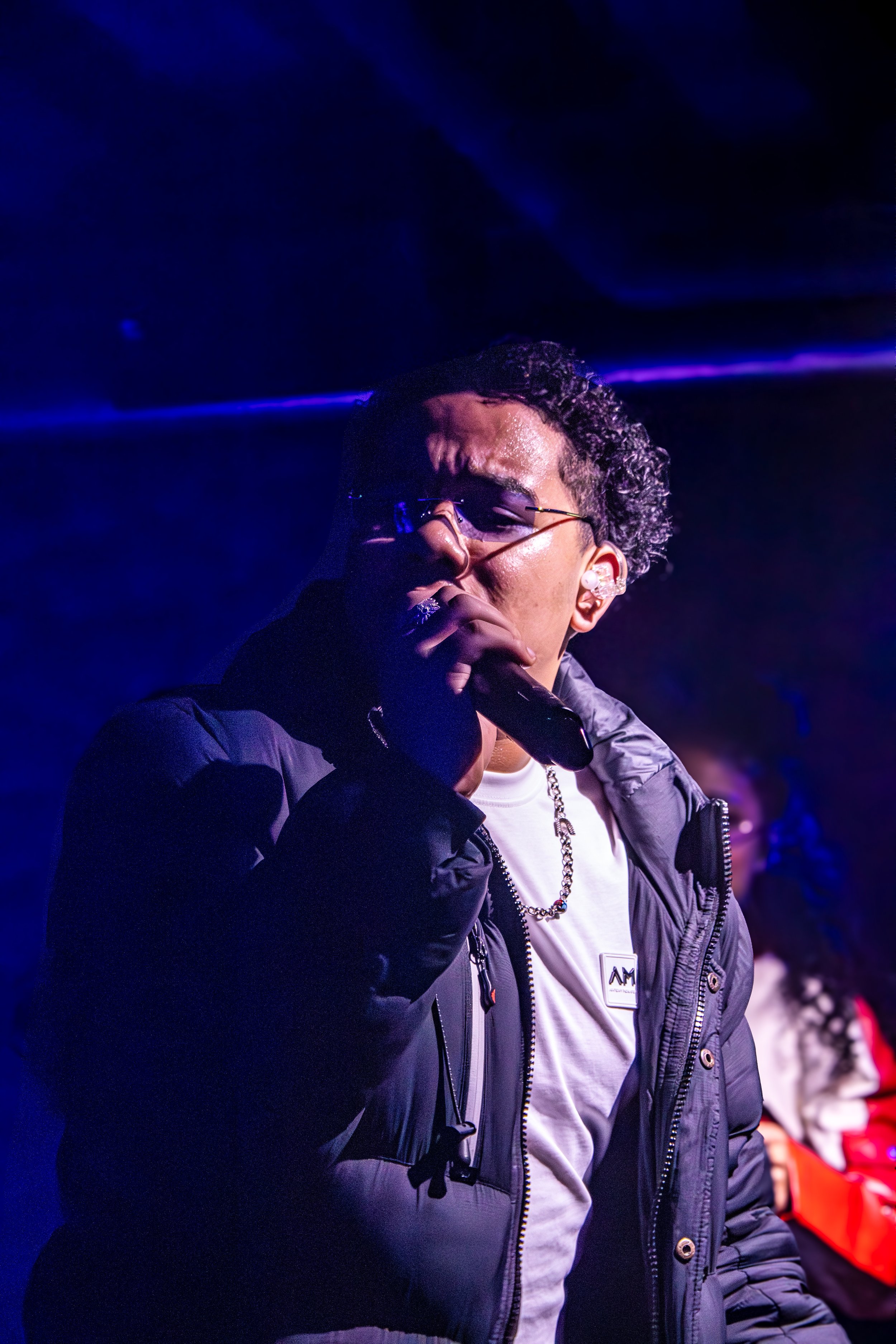A man with sunglasses and curly hair wearing a black jacket, white shirt, and silver chain, holding a microphone and performing on stage under purple lighting.