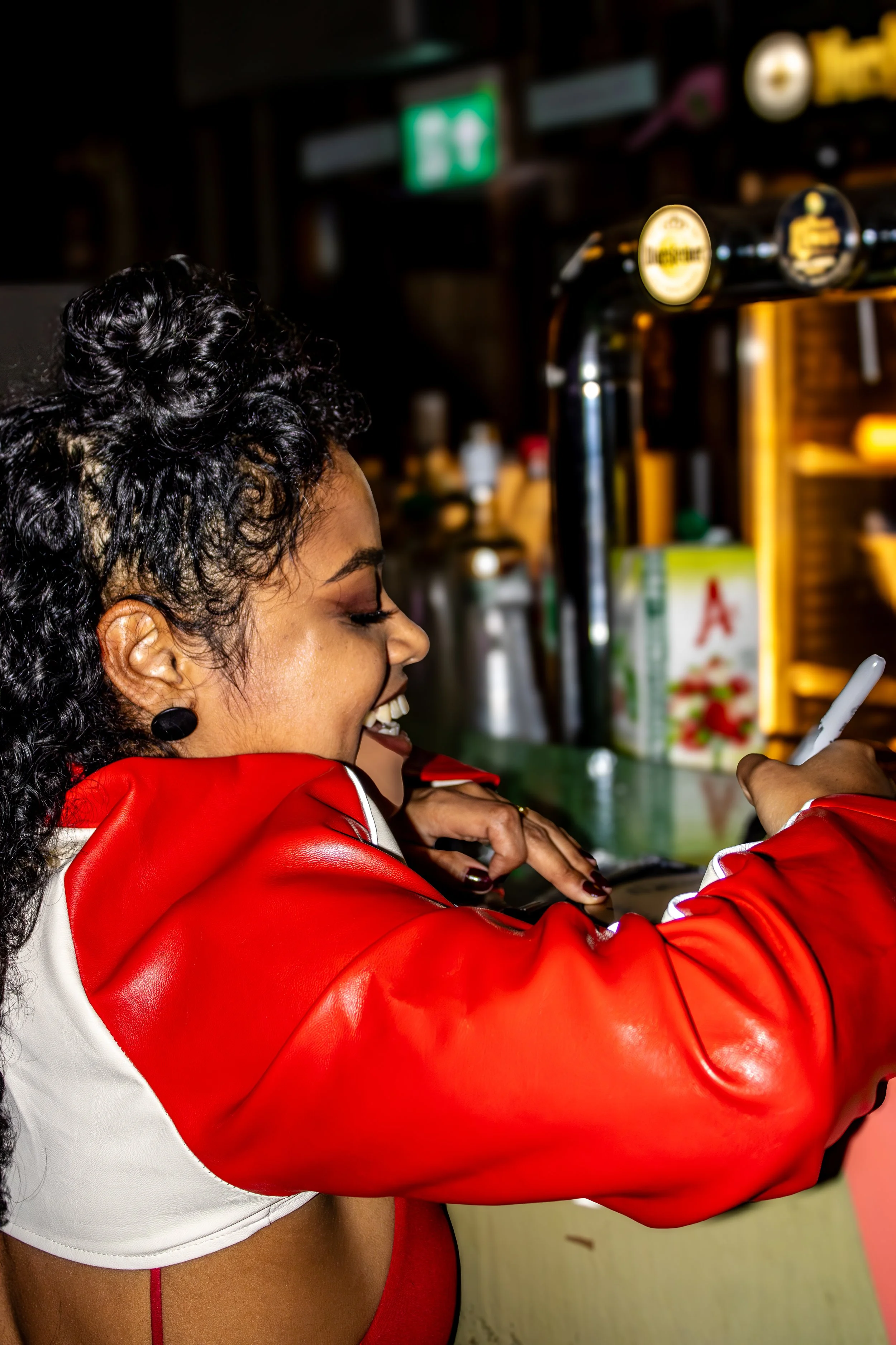 A woman with curly hair wearing a red and white jacket smiling while taking a selfie or a photo with her smartphone in a bar or restaurant setting.