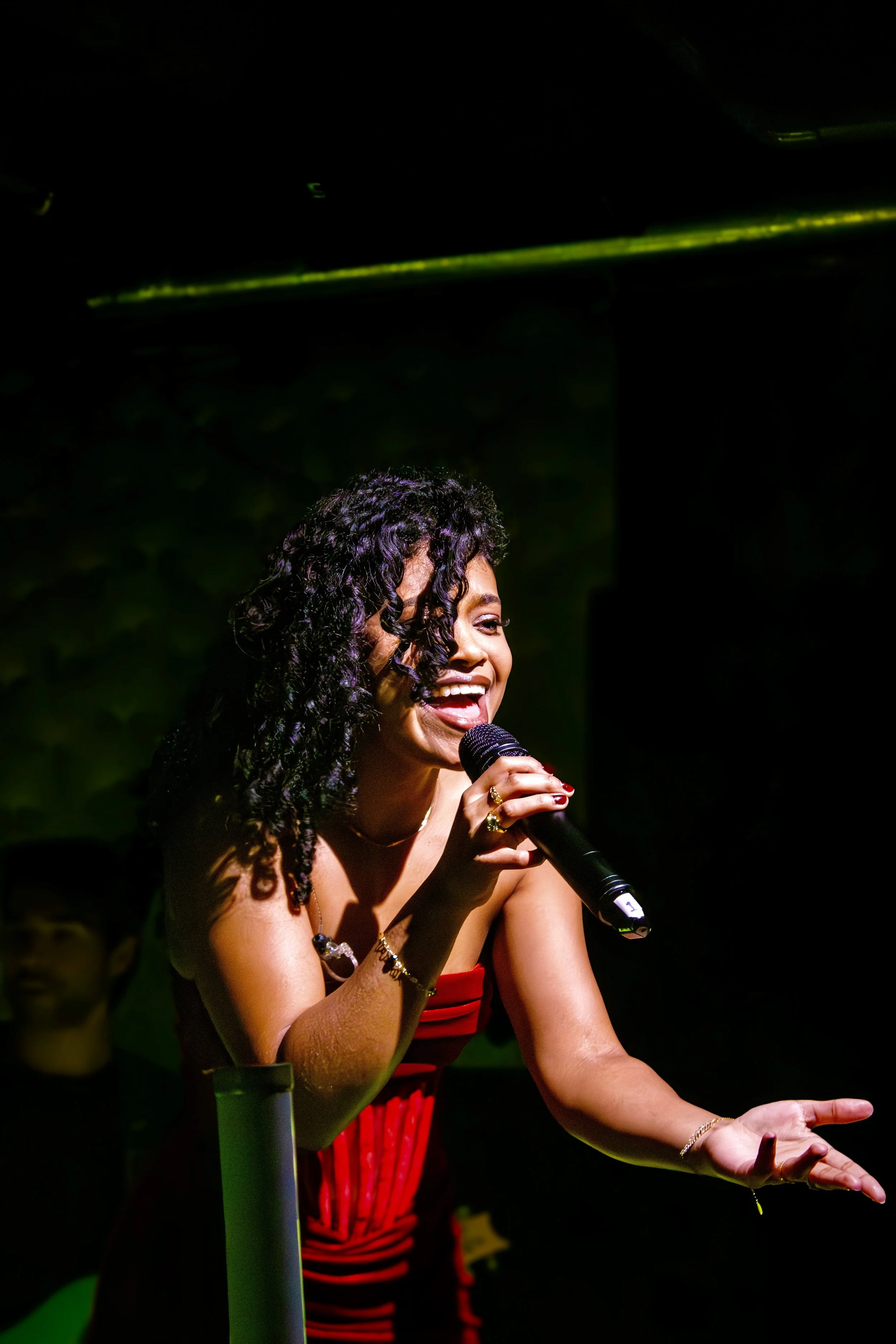A woman with dark curly hair singing or speaking into a microphone on stage, wearing a red strapless dress, surrounded by colorful stage lighting.